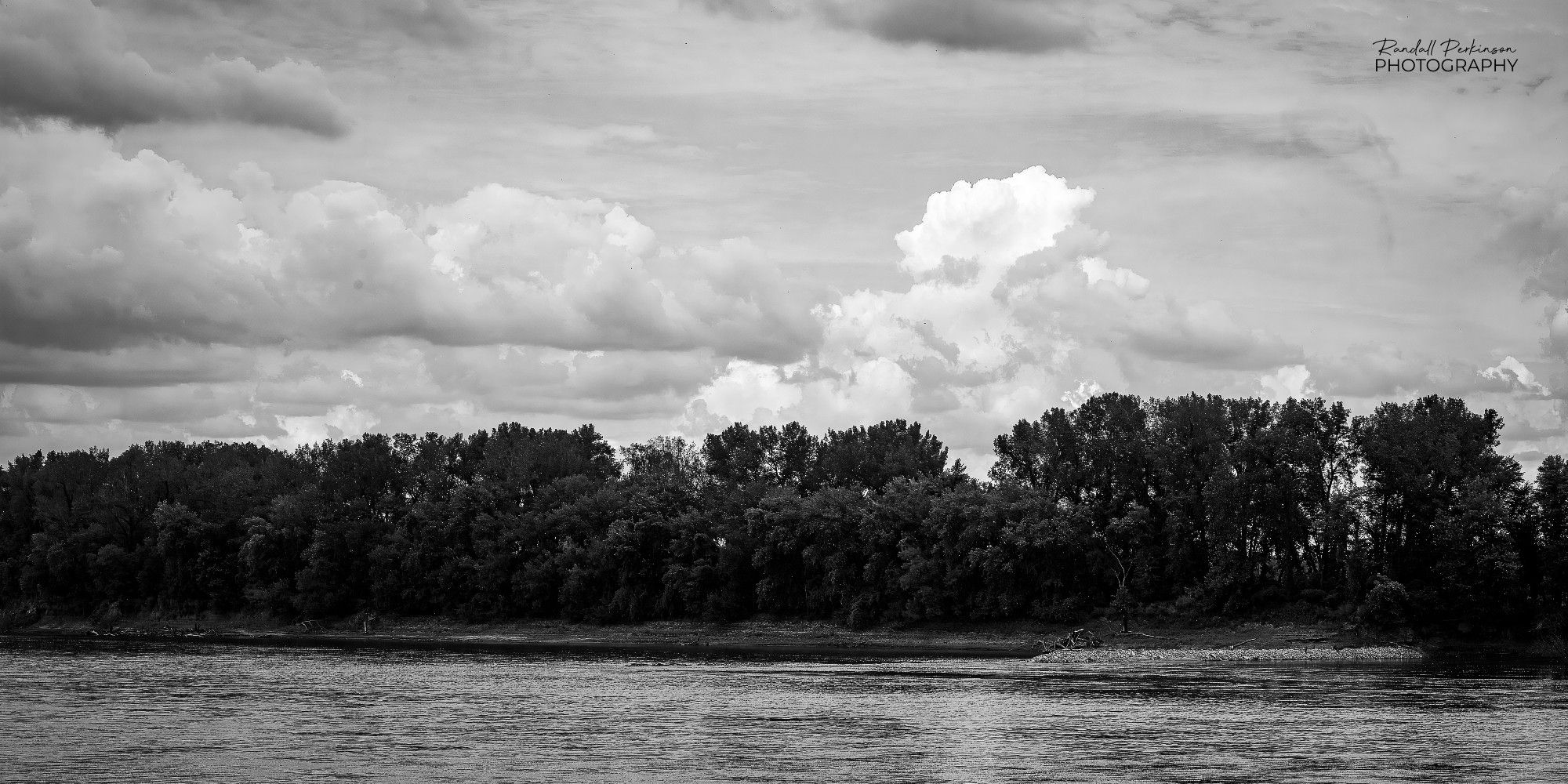 A large river flows past a wooded area with storm clouds forming above.