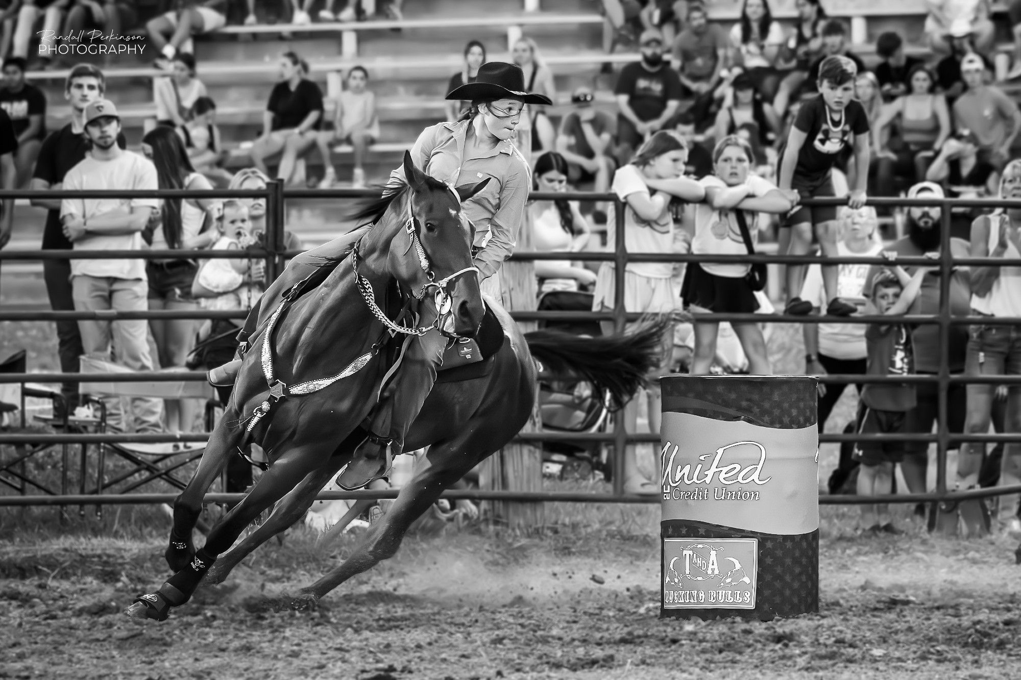 A barrelracer and her dark colored horse lean into a turn around a barrel in a competition to see who can run the course in the shortest time.