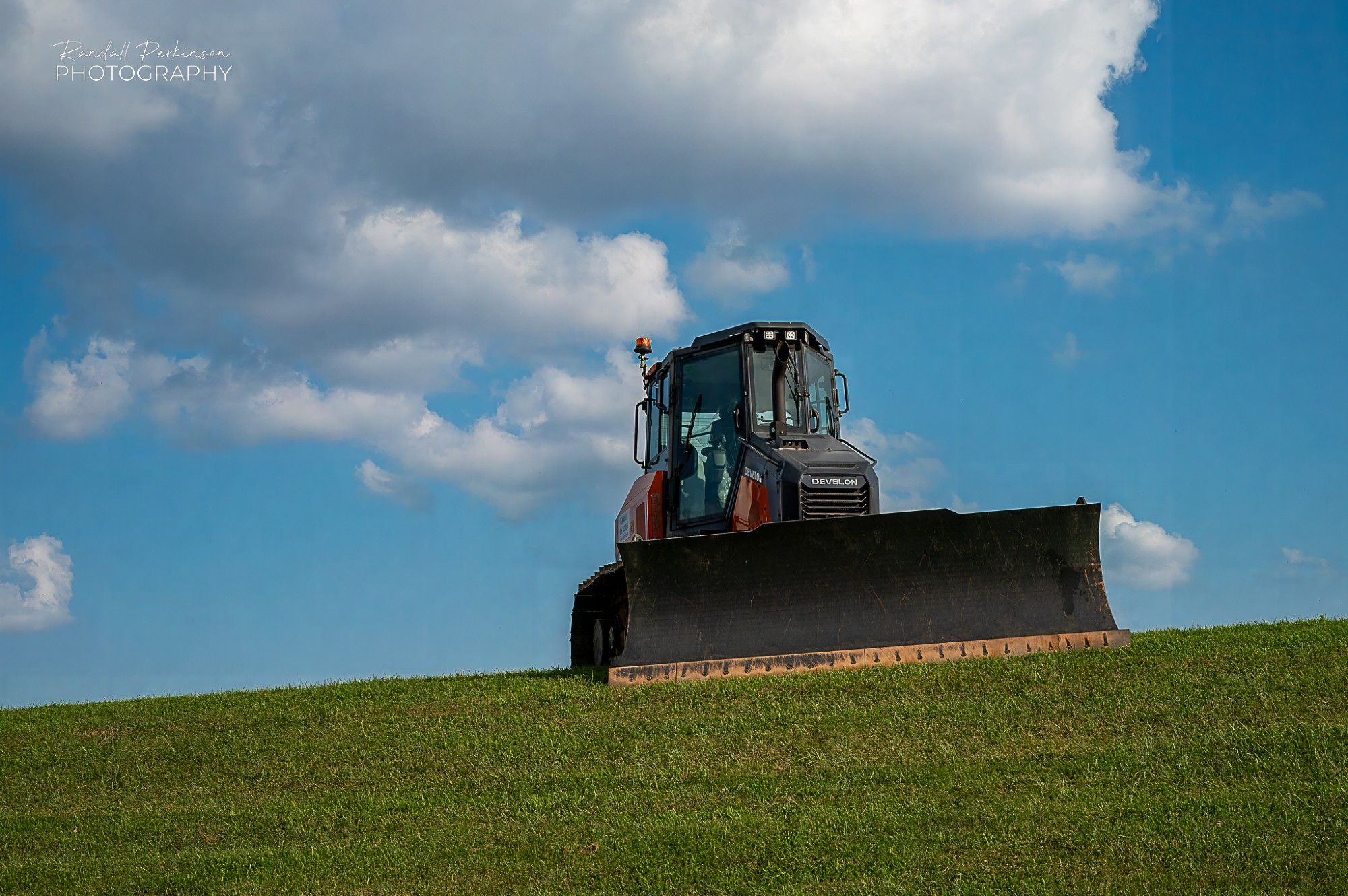 A bulldozer faces the camera at the top of a green, grass slope with blue sky and clouds in the background.