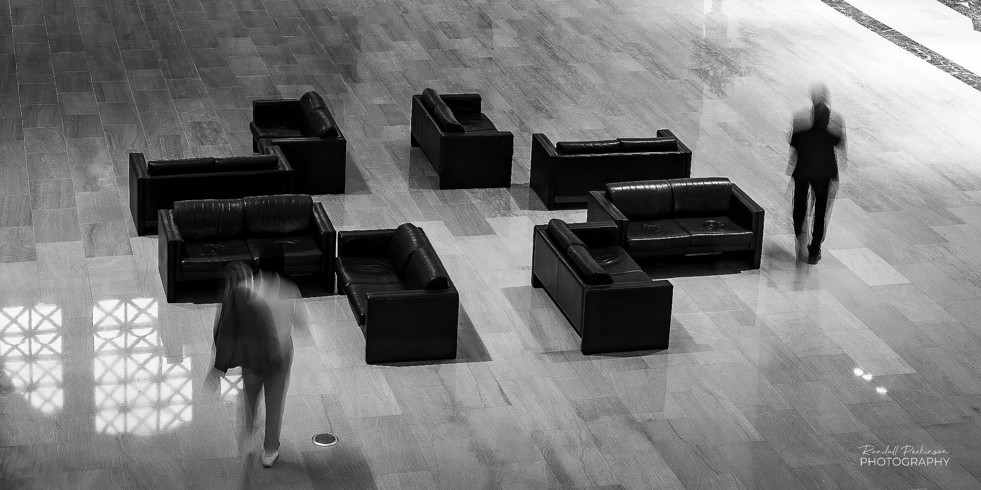 Eight black leather love seats sit in the middle of an art museum main haul with two blurred people walking past them.