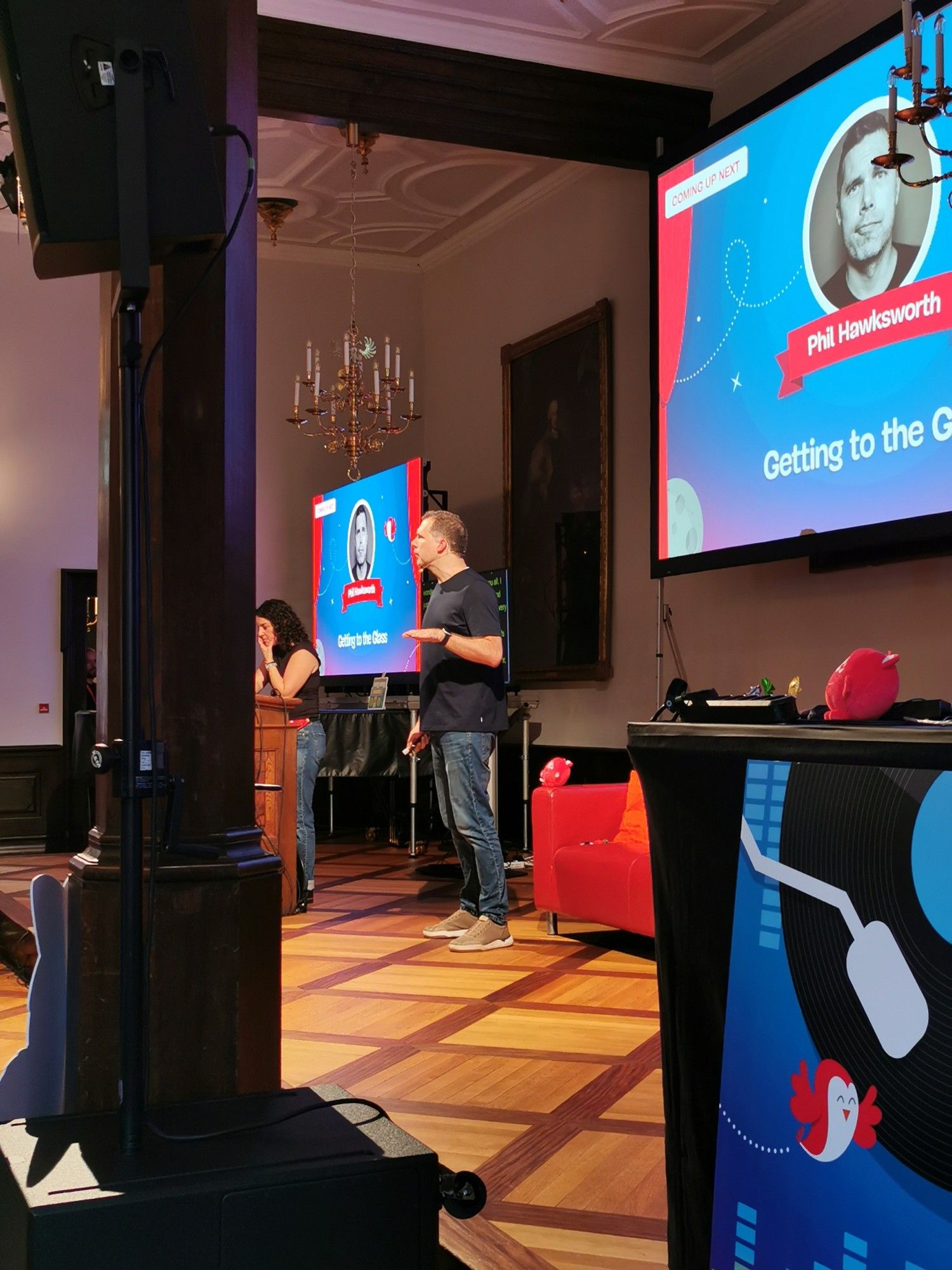 The image shows a close-up of a speaker, Phil Hawksworth, presenting at the Smashing Conference 2024 in the historic Kaufhaus building in Freiburg. He stands on a wooden floor stage, dressed casually in jeans and a t-shirt, speaking in front of a large screen displaying his name and the presentation title, "Getting to the Glass." The room's elegant decor includes chandeliers, wooden pillars, and classic artwork on the walls. Another person, possibly an event staff member, is seen behind a podium on the stage. The setting appears professional and well-organized, reflecting a vibrant conference atmosphere.