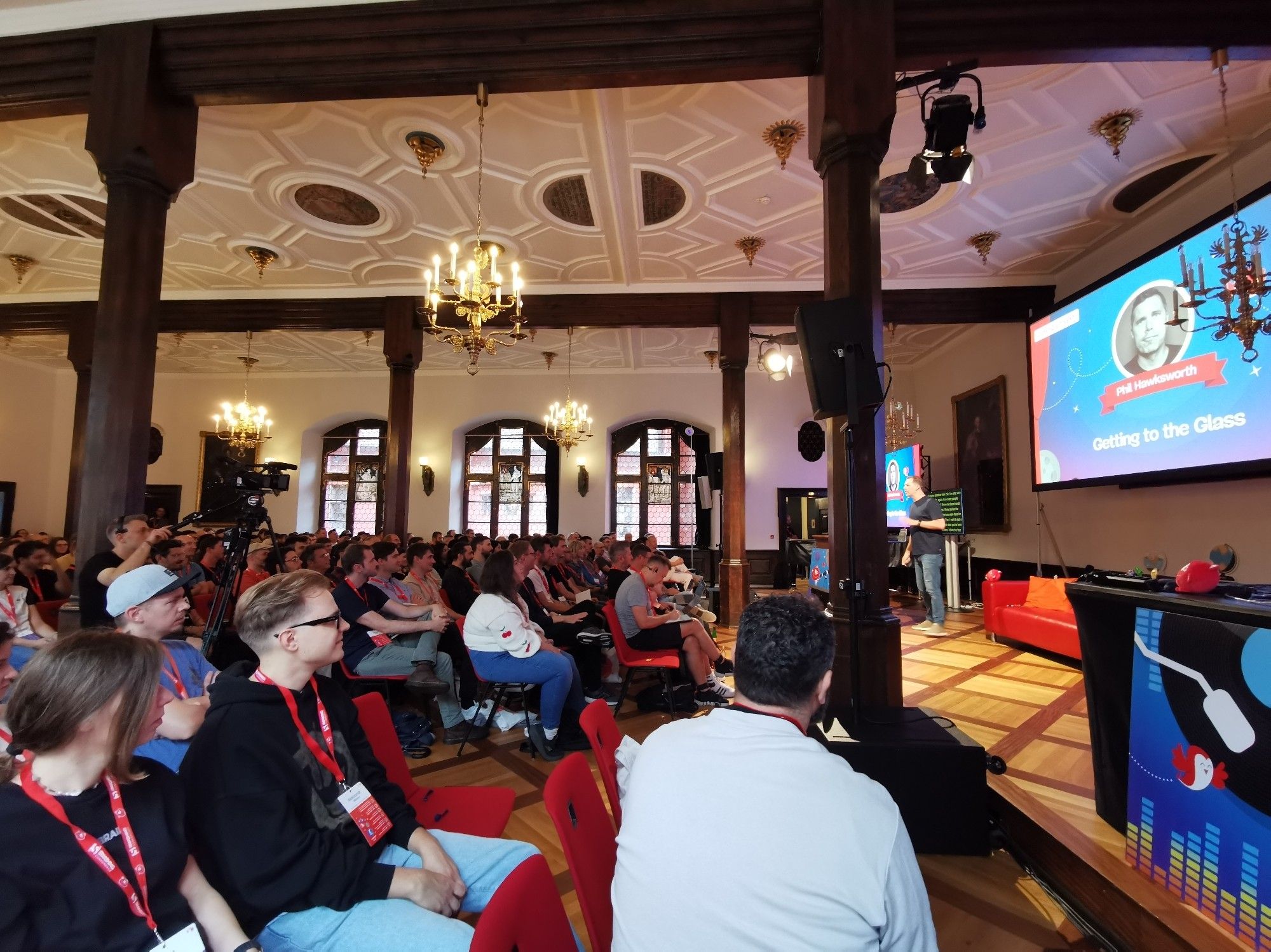 The image shows a large, ornate room filled with attendees seated in rows, listening to a presentation at the Smashing Conference 2024, held in the historic Kaufhaus building in Freiburg. The room features high ceilings with intricate designs, large windows, chandeliers, and wooden beams. On the right side, a speaker is presenting on stage with a screen displaying the presentation title "Getting to the Glass" and the speaker's name, Phil Hawksworth. Attendees are engaged, with many wearing conference lanyards and badges. The atmosphere appears professional and focused.