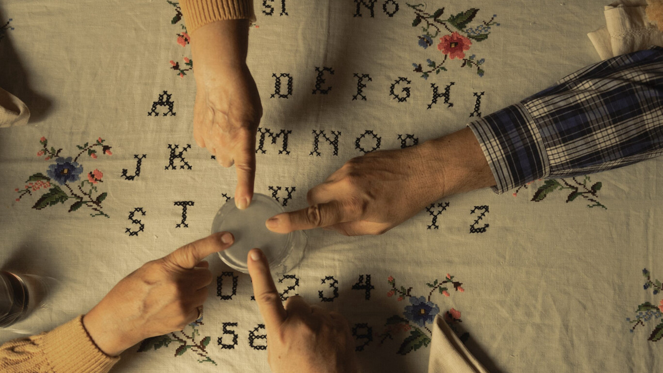 Cuatro manos con su dedo índice apoyado en un vaso. Están haciendo ouija, pero el tablero es un mantel bordado.