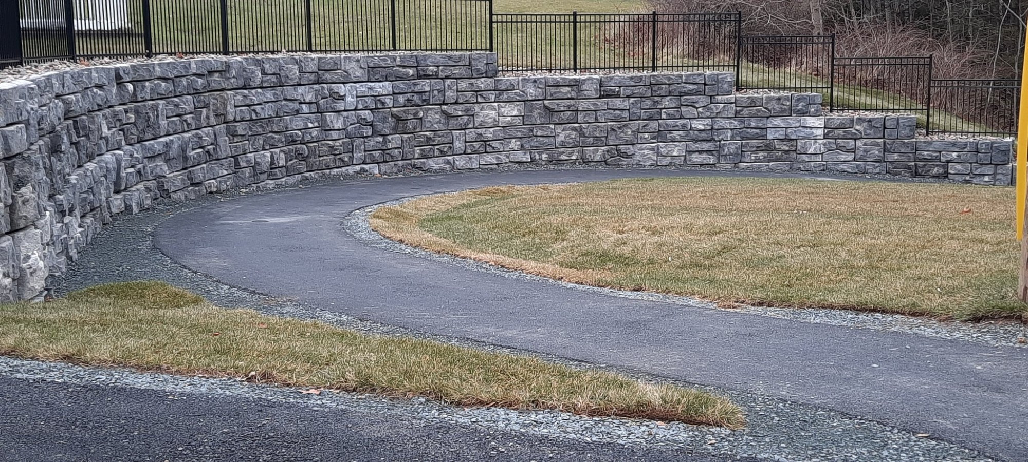 A concrete wall wraps one side of a sloping asphalt connector trail. An iron fence sits atop of the wall.