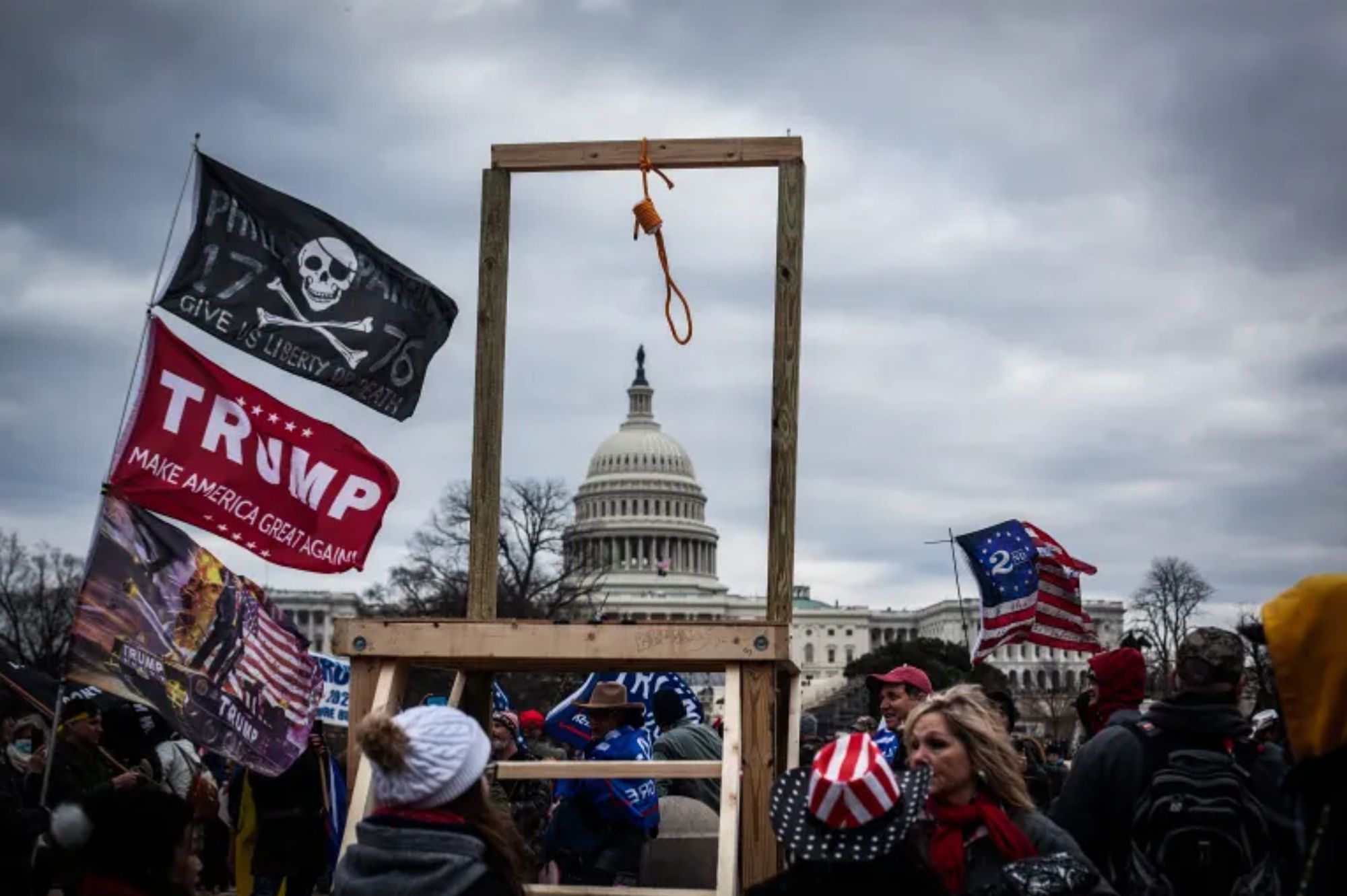 Photo of the infamous improvised gallows constructed outside the Capitol building by rioters on January 6th. The gallows frames the Capitol dome in the background, and rioters fly Trump flags and congregate nearby.