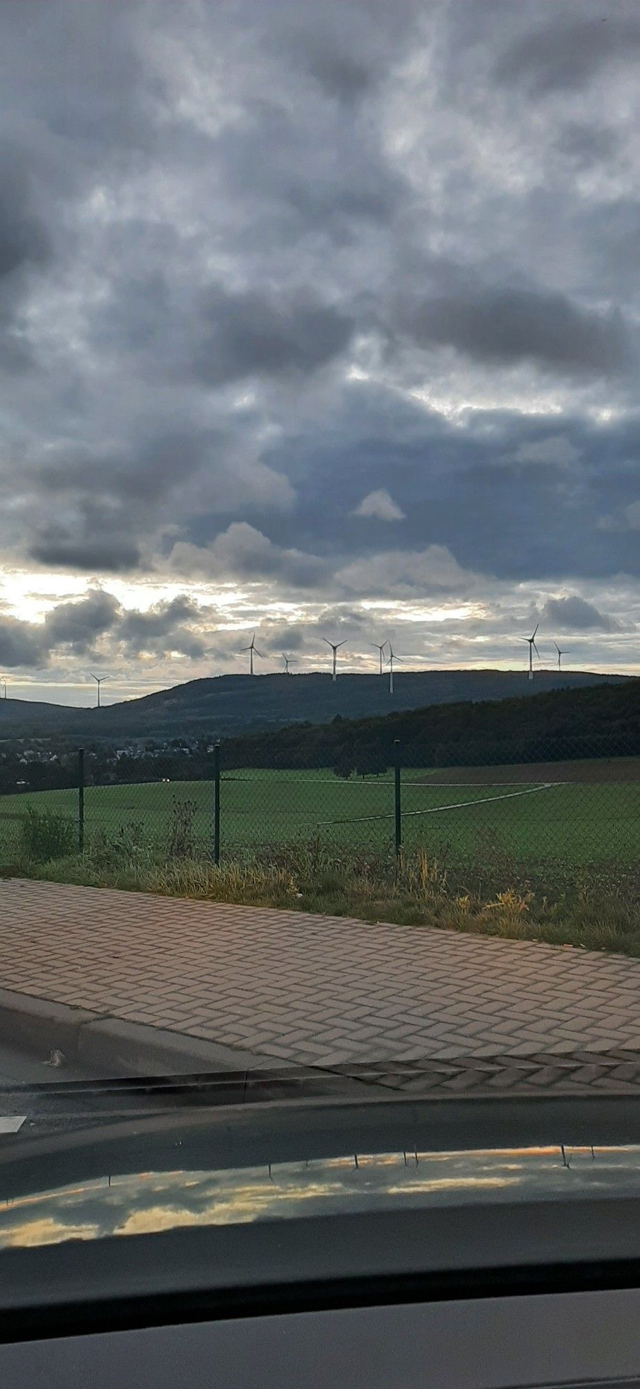 Ausblick vom Rastplatz Ellern an der B50 Richtung Mainz.
Zu sehen sind Graue Wolken mit leicht orangenen Touch von der aufgehenden Sonne. Bewaldete Berge mit Windrädern, eine Ortschaft und grüne Wiesen.