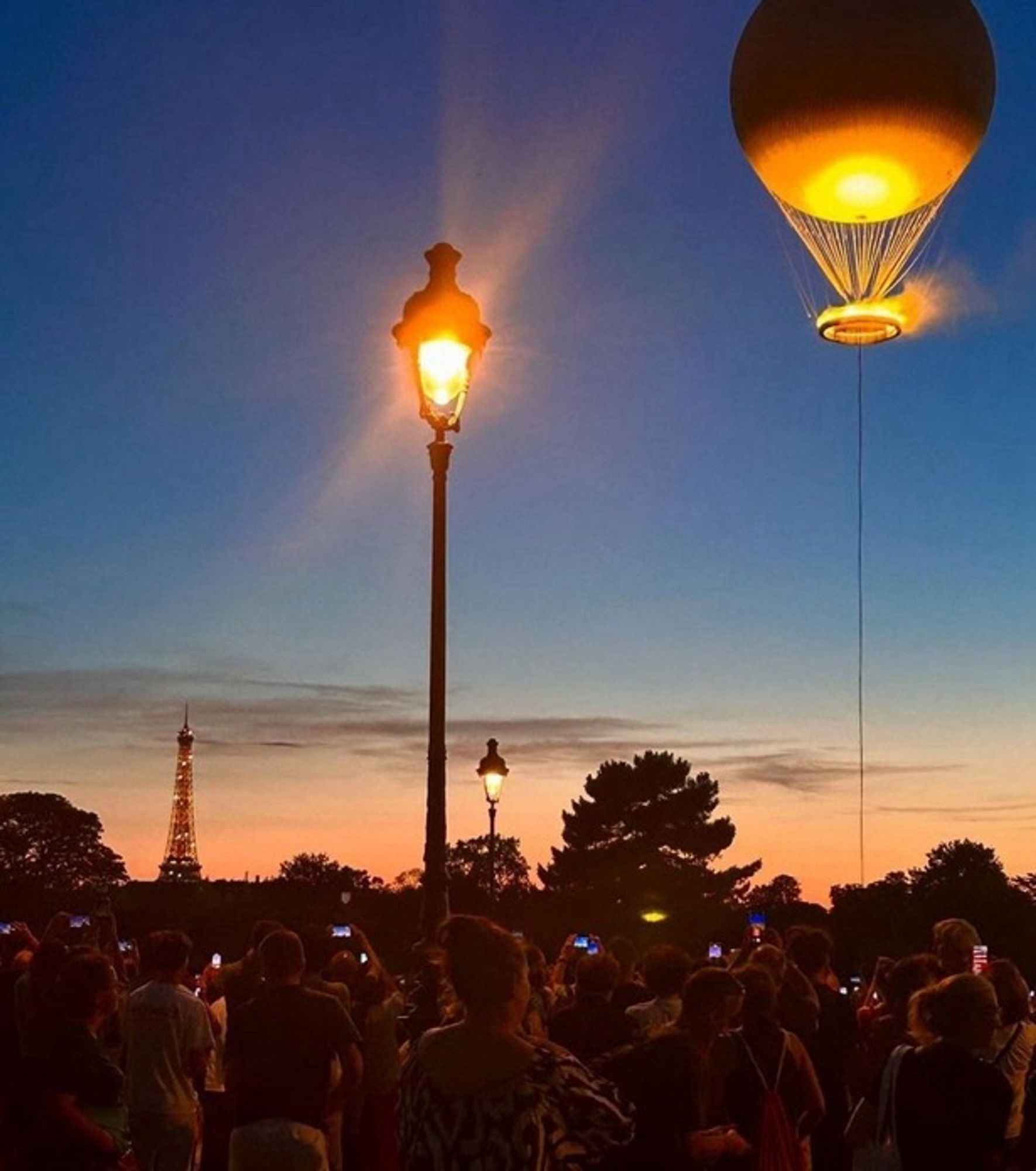 Vasque dans le ciel de Paris au crépuscule. En arrière-plan le haut de la tour Eiffel. Au premier plan, le public venu l’admirer, téléphone à la main pour immortaliser le moment