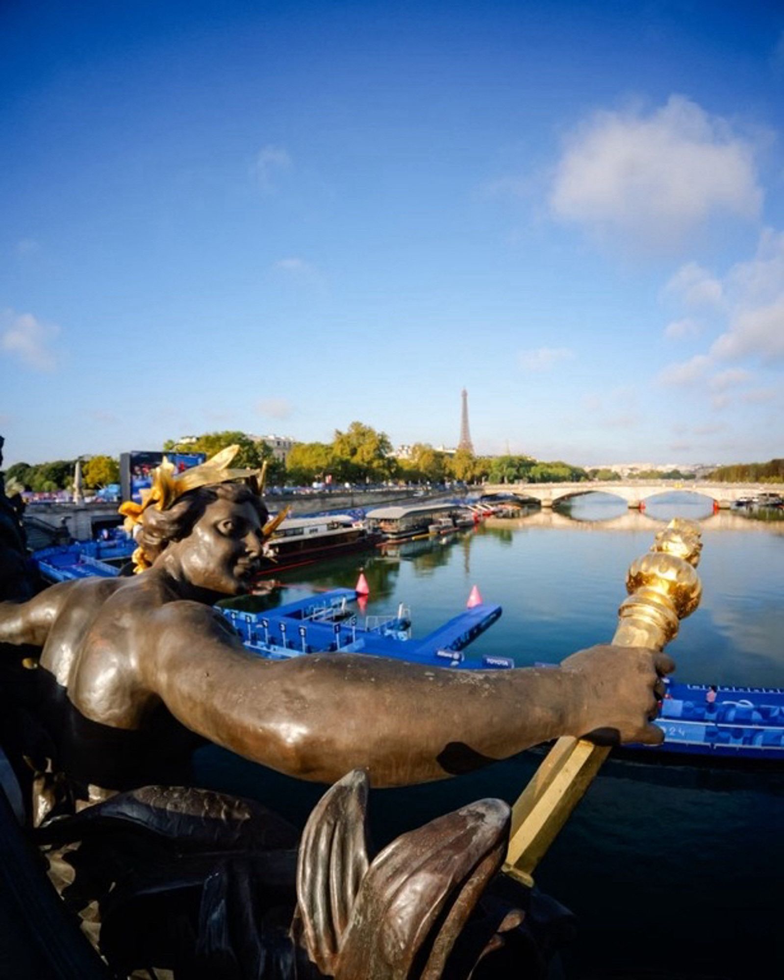 Vue du Pont Alexandre III sur le ponton de départ du para triathlon avec en premier plan une des statues d’ornement du pont et toute petite en arrière-plan, la tour Eiffel