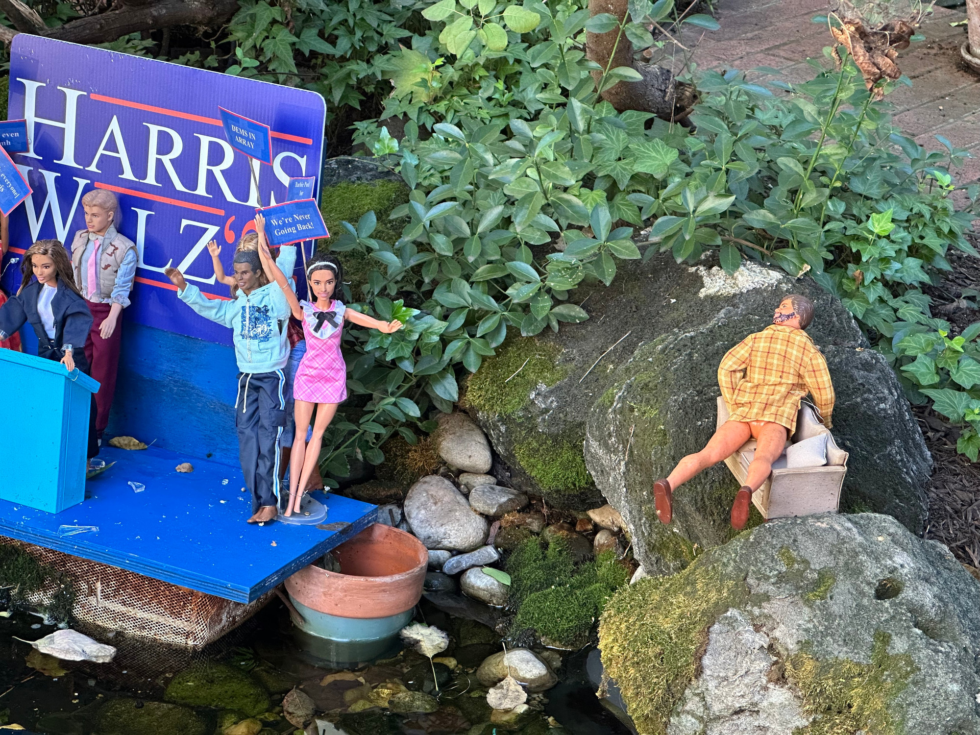 Display of Barbies on HarrisWalz campaign stage with JD Vance looking on from the side while fucking a couch with no pants on. 