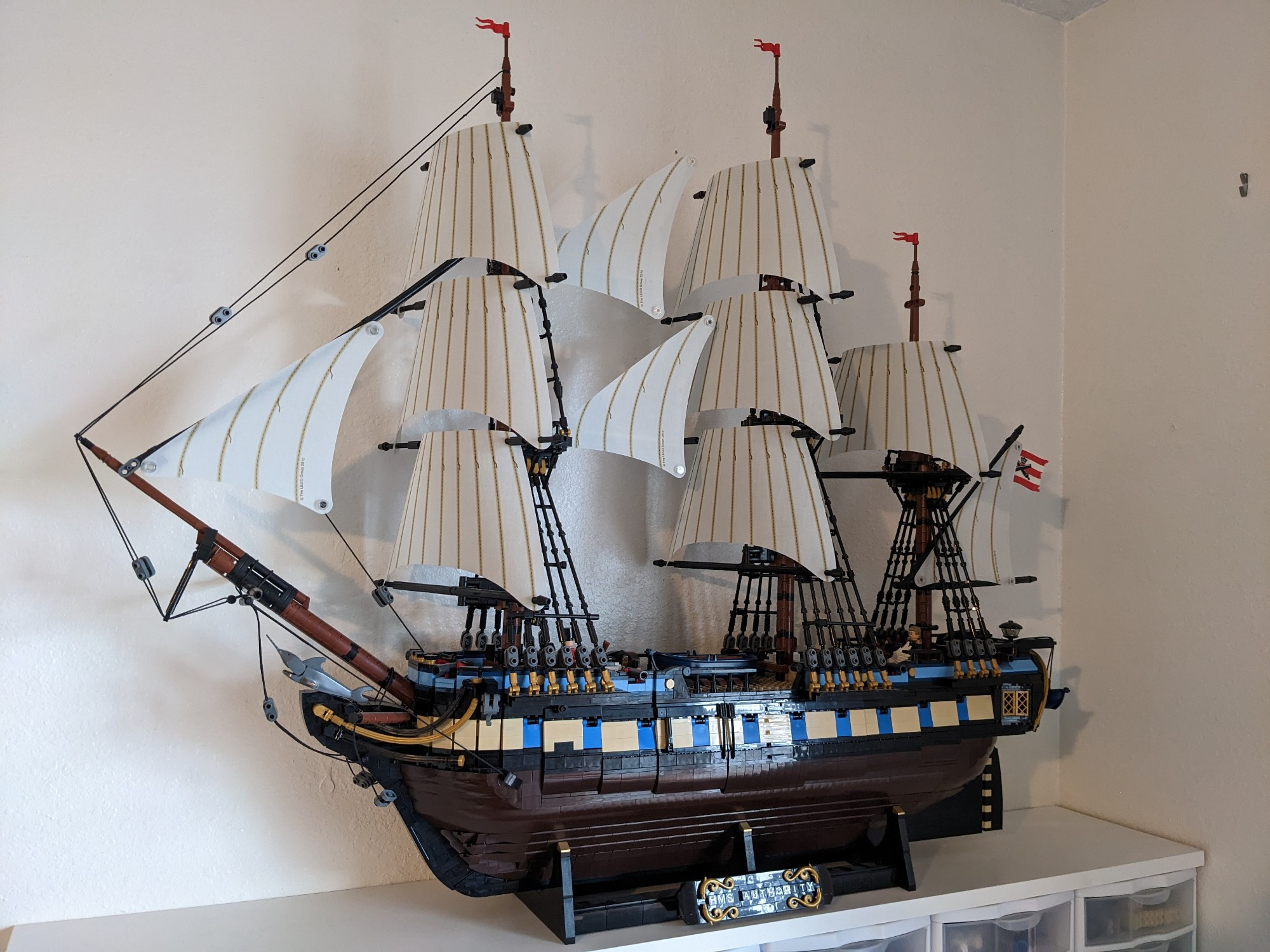 A custom LEGO model of a three-masted sailing ship. The ship is held on a black stand bearing the name "HMS Authority". The hull is a dark brown, and there are stripes of tan and blue mixed in with black. The sails are an off-white, and rigging is represented with many things black pieces and some string. The model is large, and the entire thing rests up on a display shelf.