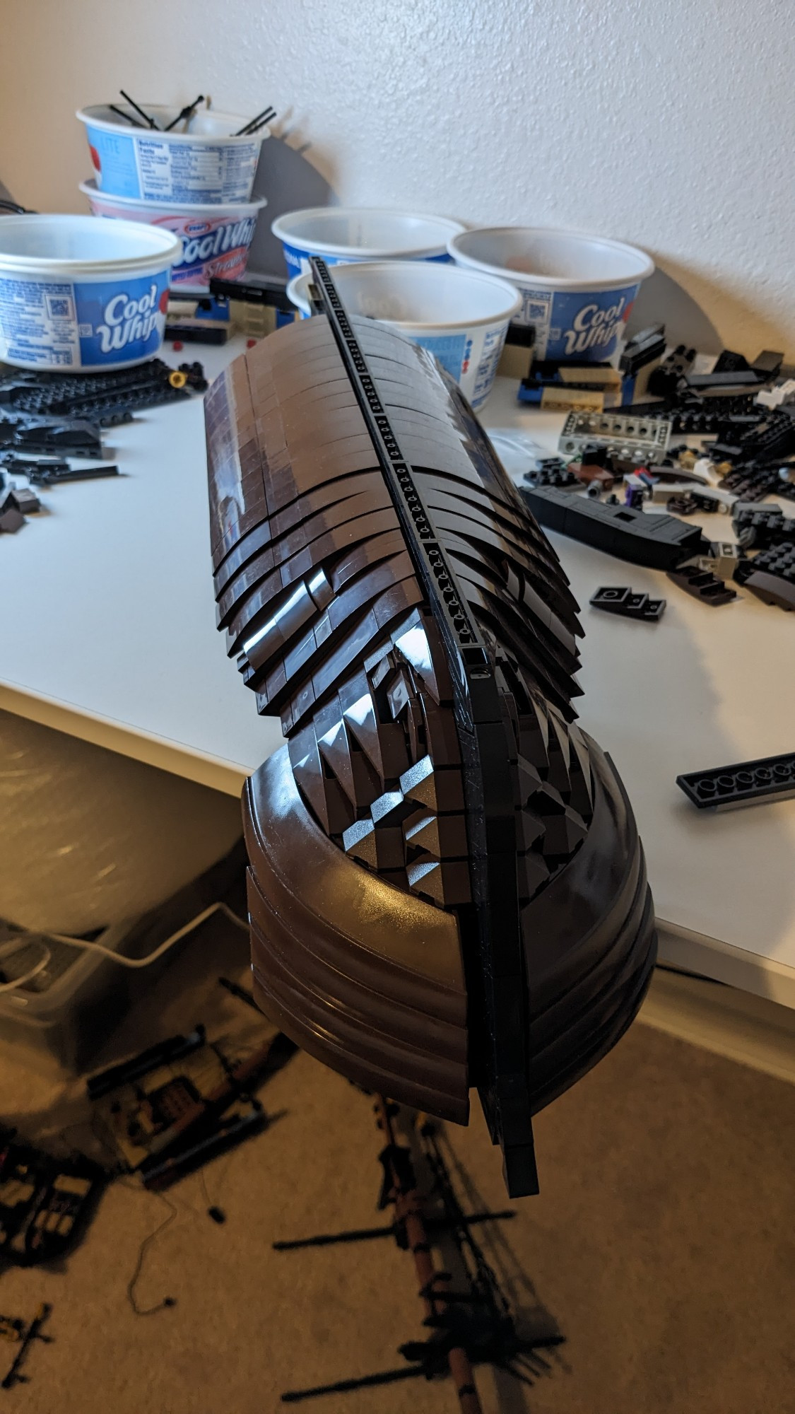 An alternate view of the work-in-progress LEGO model, this time flipped upside down. This shows off the rounded shaping of the hull. It rests on a white table that is covered in other LEGO pieces, and some repurposed containers.