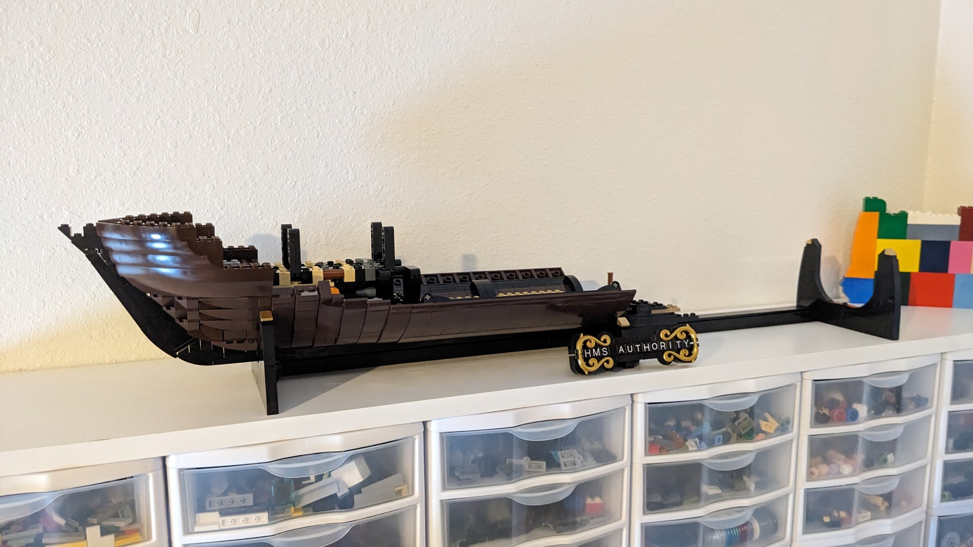 A work-in-progress LEGO model of a sailing ship. A black display stand rests on a shelf, and includes a nameplate adorned in gold detailing that reads, "HMS Authority." On the stand is the beginning of the ship, which features lower portions of the hull and some of the internal structure.