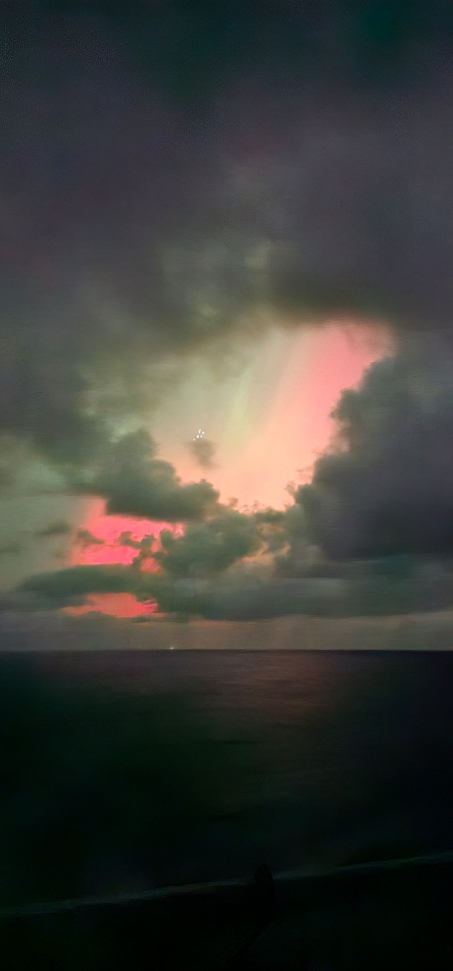 A cloudy night sky, with chunks of red, pink, and green showing through the gaps. In the centre of the picture, Ursa Minor is visible against a greyish-green sky. There's a lighthouse visible on the horizon, but I forgot to note which one.