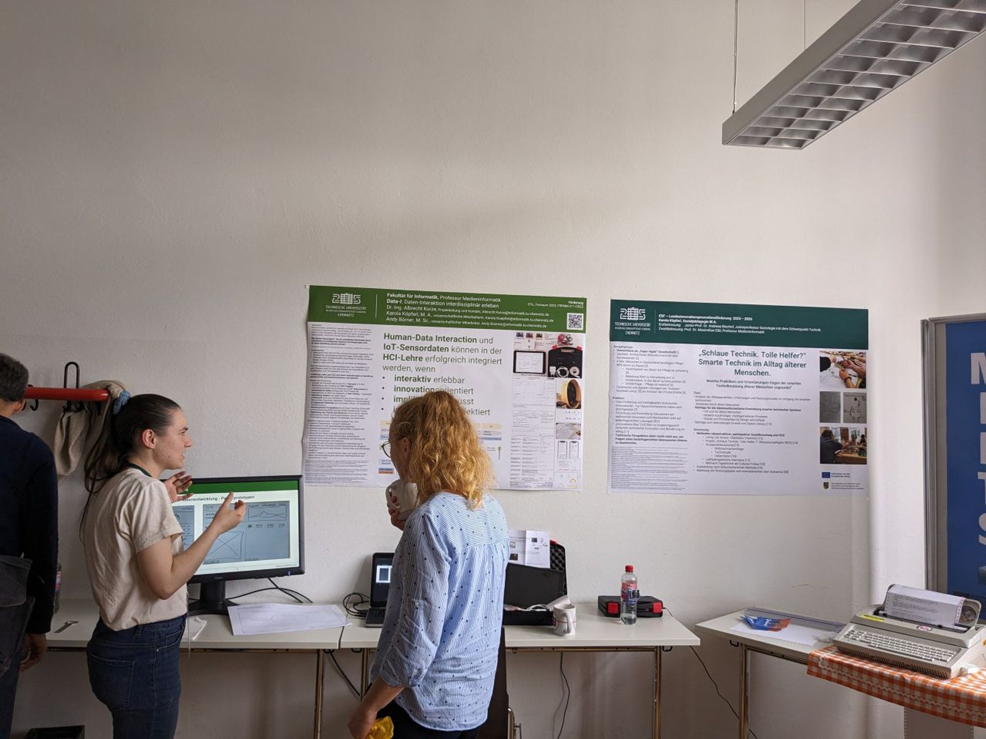 Links steht eine junge Frau und erklärt das Lehr-Lernprojekt Data-I. Hinter ihr ist ein Monitor zu sehen. Rechts hängen zwei Poster und davor steht eine Frau die den Ausführungen zuhört.