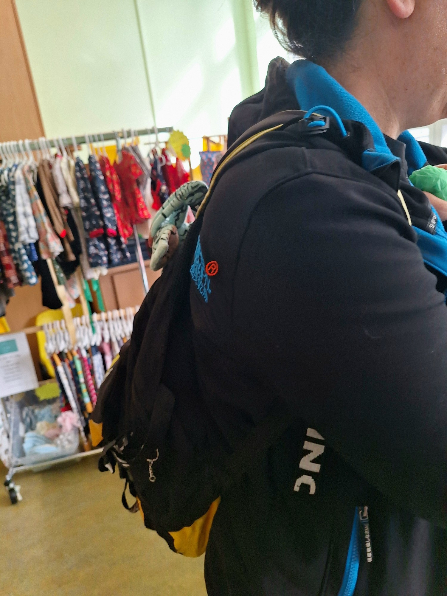 A woman standing in front of a rack of clothes. A banshee plush hangs from her bag without her knowledge.