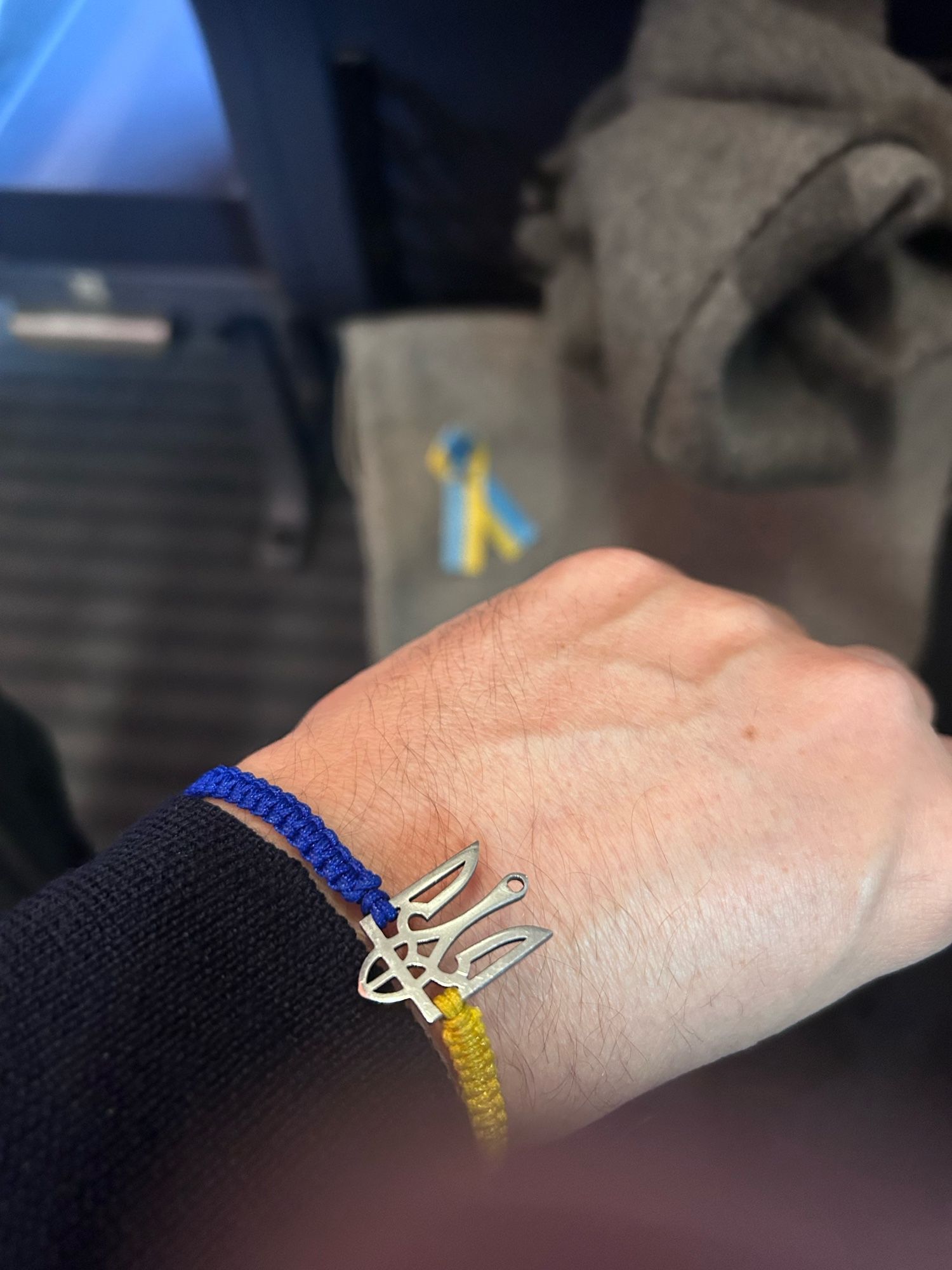 Foto meines Unterarmes mit Armbändchen in gelb und blau mit ukrainischem Wappen und im Hintergrund mein Rucksack mit einer Schleife in blau-gelb.