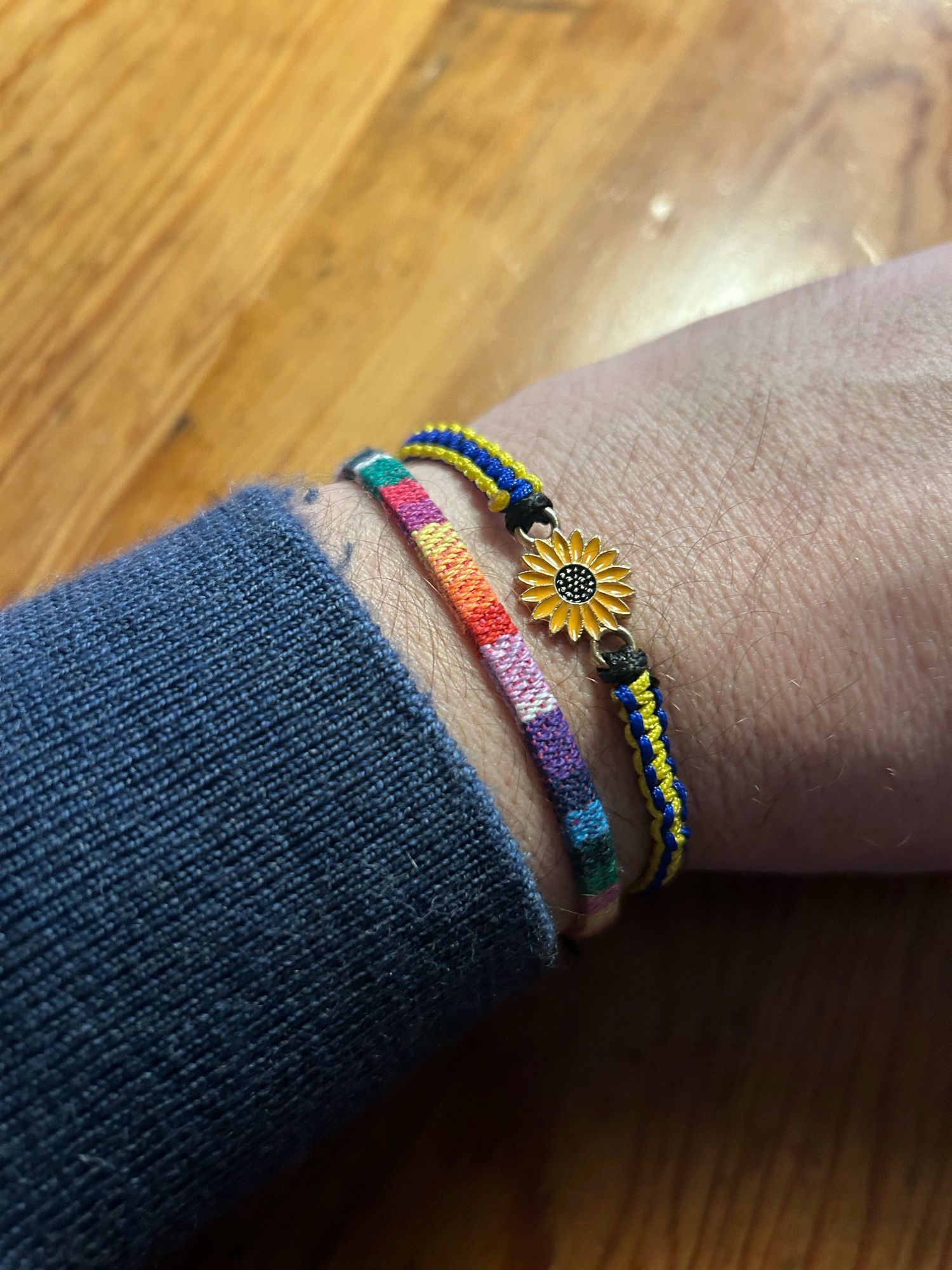 Unterarm Foto mit einem Armbändchen in Regenbogenfarben und einem Armbändchen in blau-gelb und einer kleinen Sonnenblume