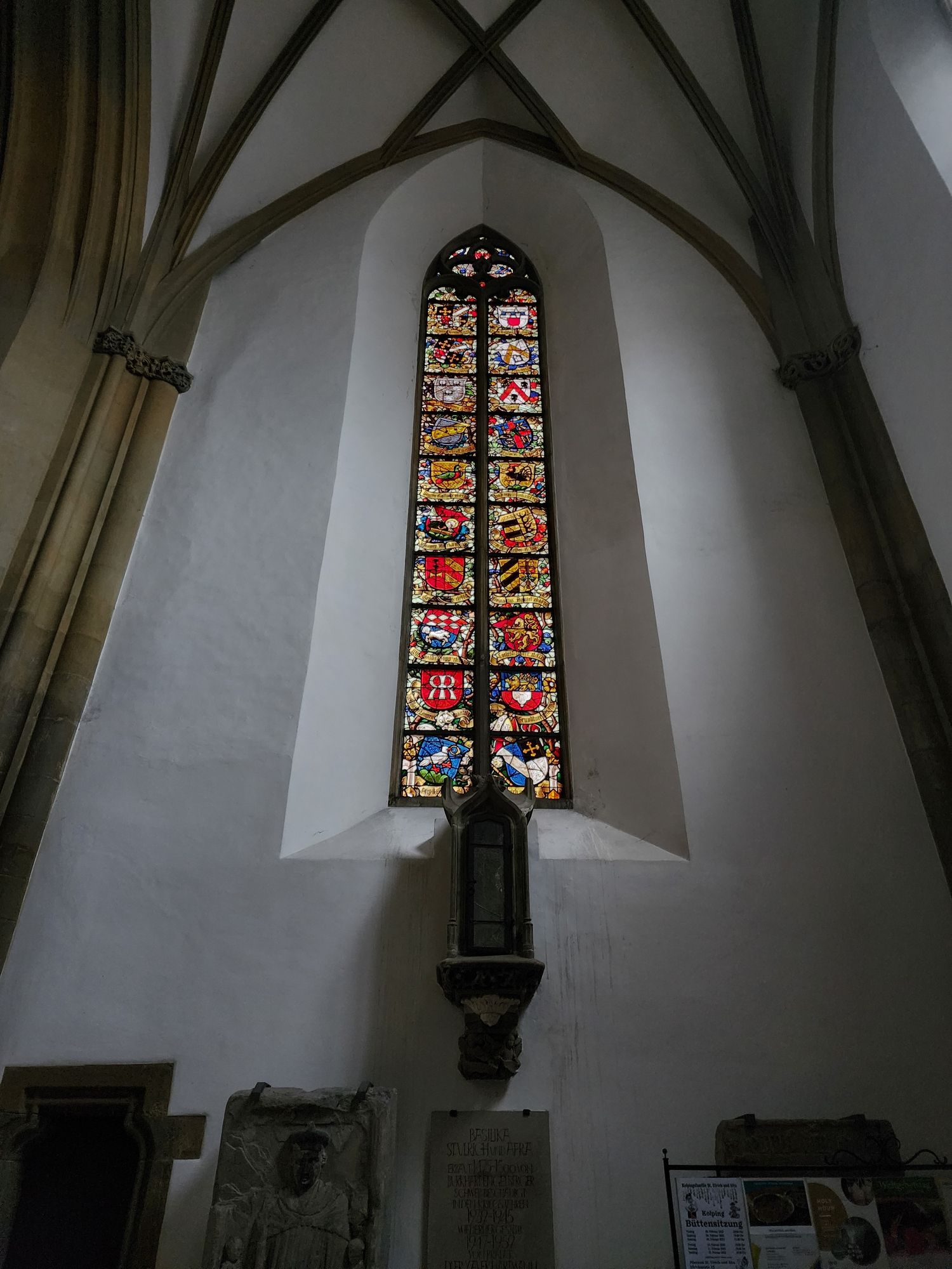 Tall, narrow stained glass window deep inset in a white wall with vaulting and brown ribbing. The window is composed of two columns of ten squares, each of which contains a very colorful coat of arms.