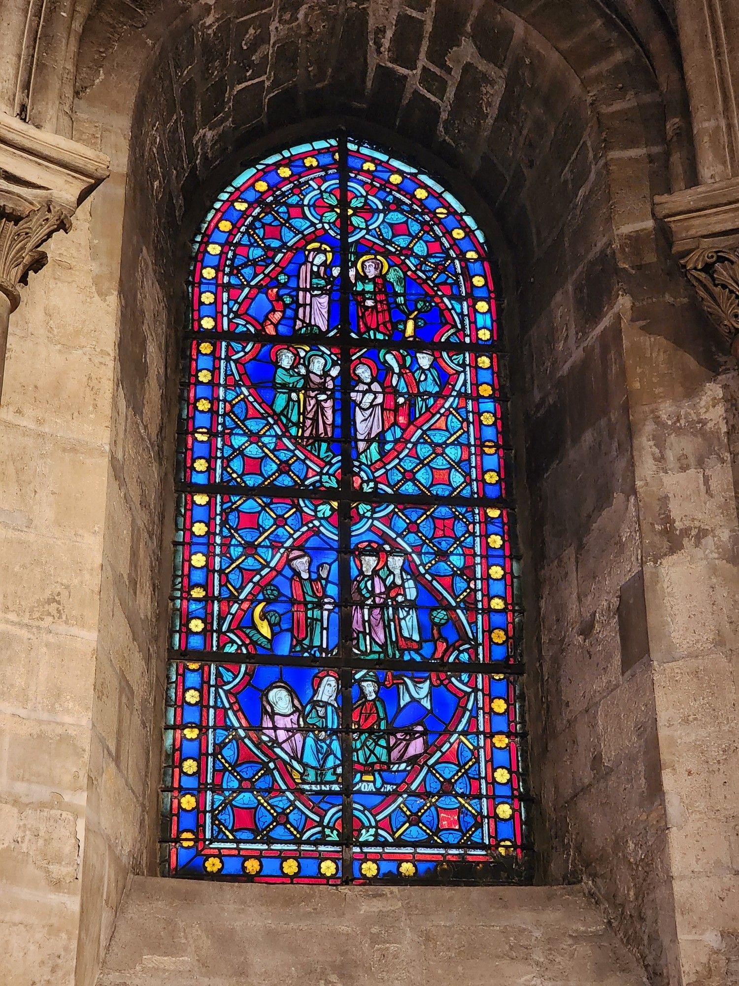 tall, arched stained glass window of mostly blue and red tones. it is divided by a metal grating into four rectangular sections, each of which depicts a scene from the gospels or the saint's vita.