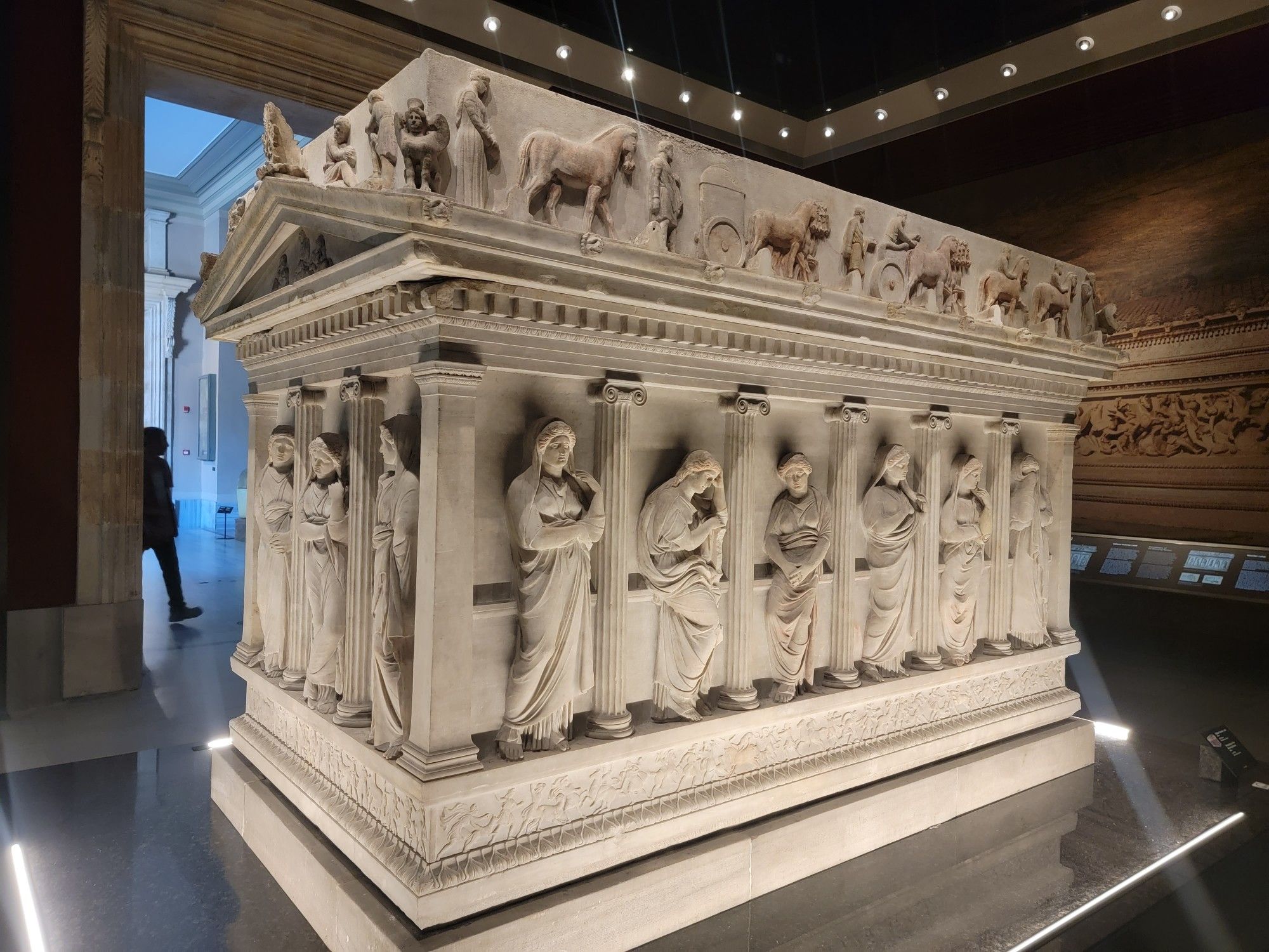 Large rectangular marble sarcophagus in the shape of a Greek temple, like the Parthenon. Between each of the pillars is a female figure in a veil. Along the very top are horses and chariots in motion.