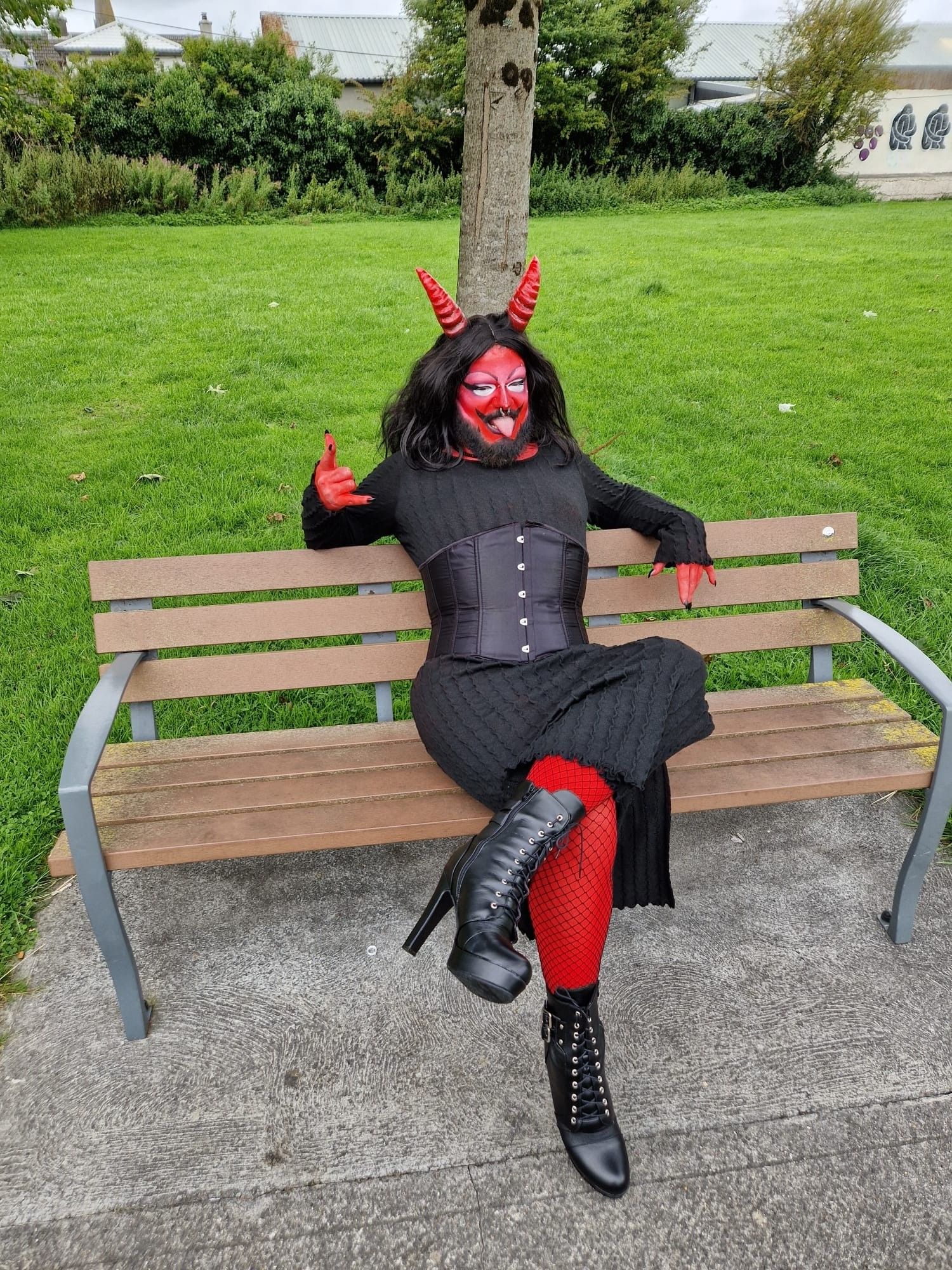 Me in full red demon drag in a black dress and corset and heels sticking my tongue out and giving the devil horns sign with my hands sitting on a park bench