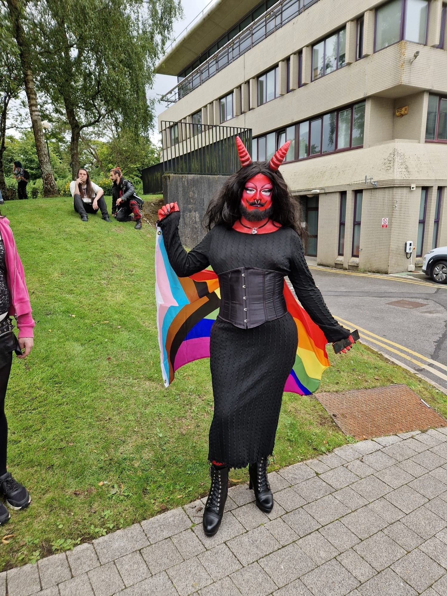 Me in full demon drag with a progress pride flag in a black dress and corset