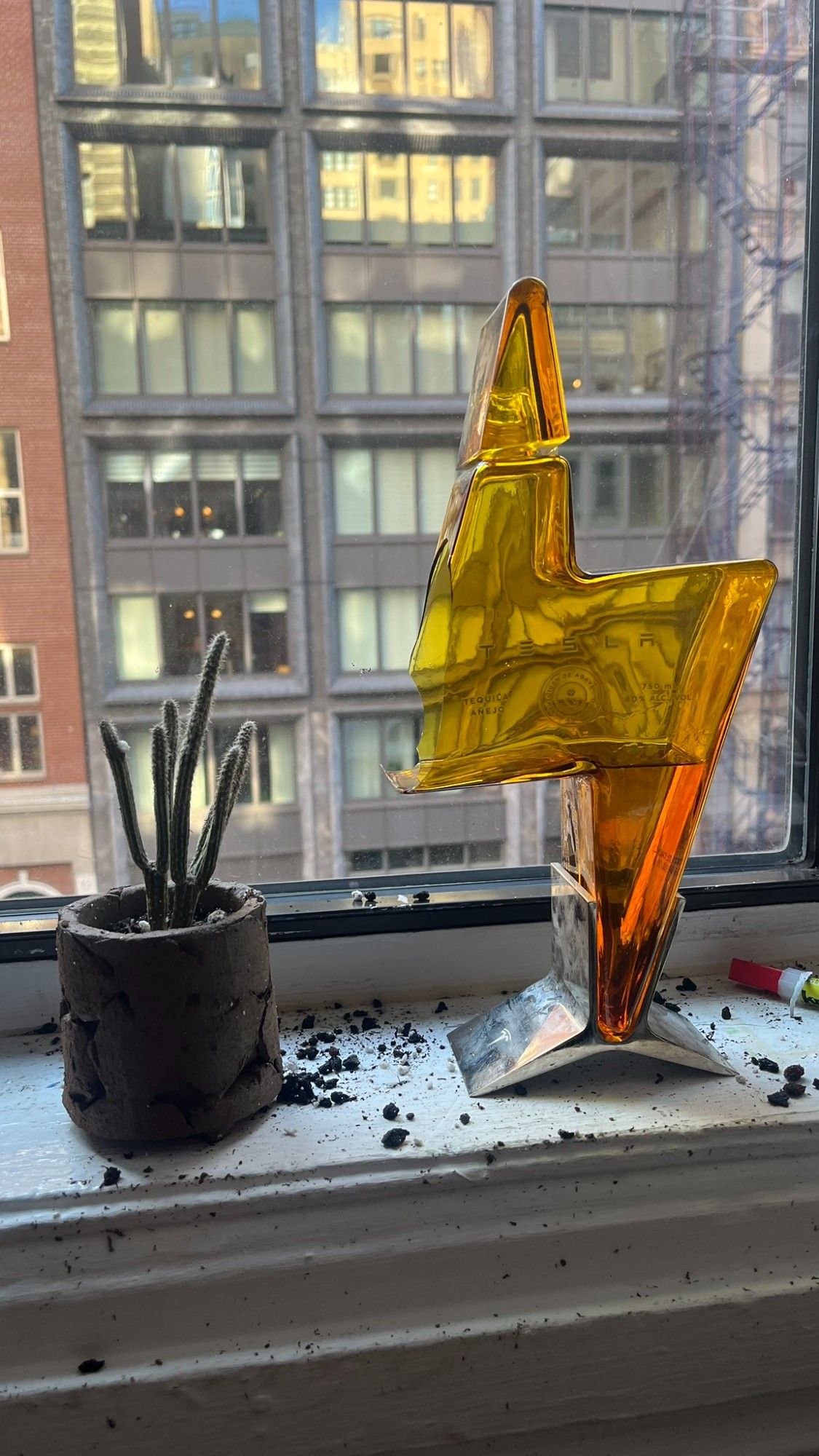 A lightning-bolt-shaped bottle of Tesla-branded tequila, standing on a windowsill with a massive exploded hole around the downward-facing suspended corner of the design.