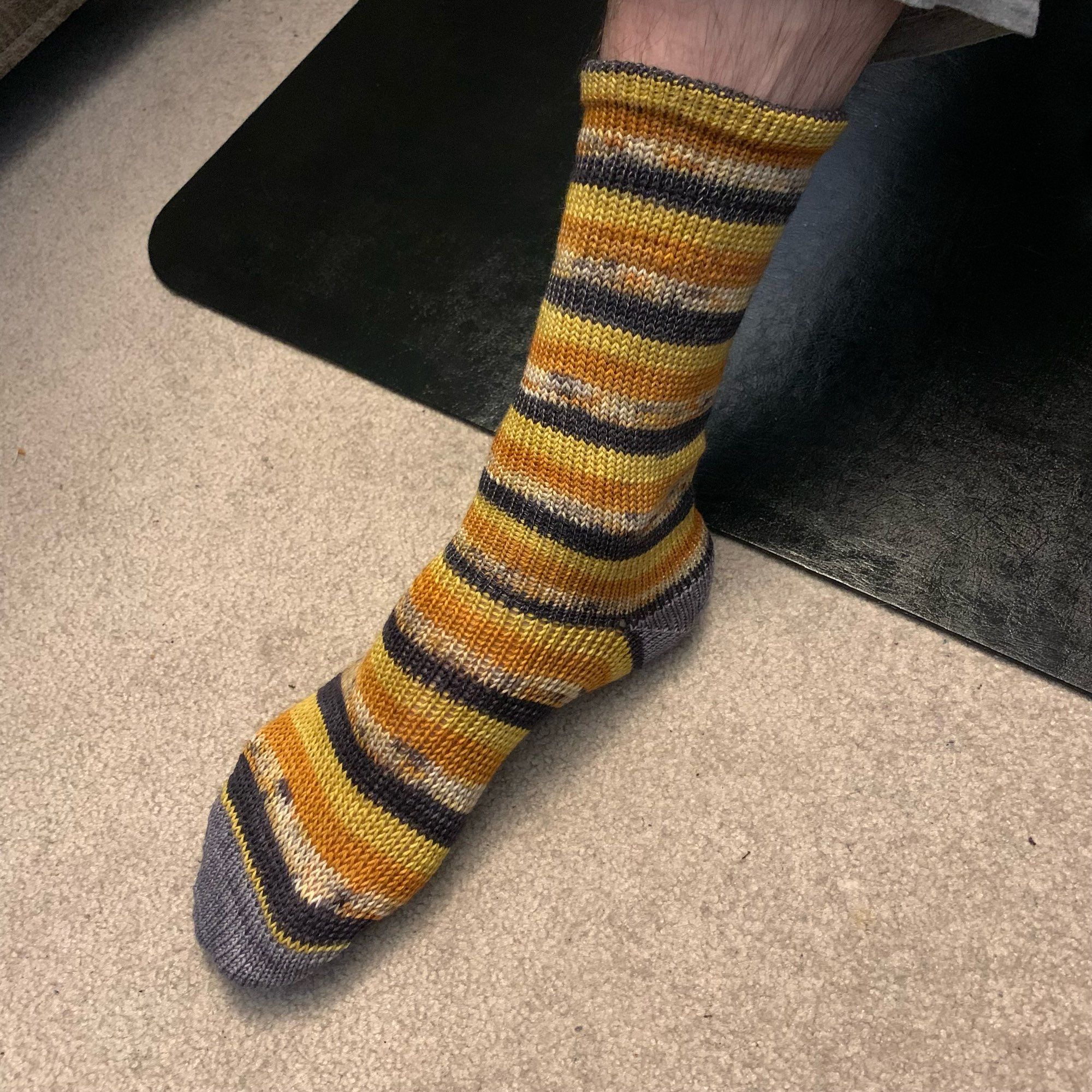 An orange, yellow, black, and variegated knitted Halloween sock with grey toes and heel.