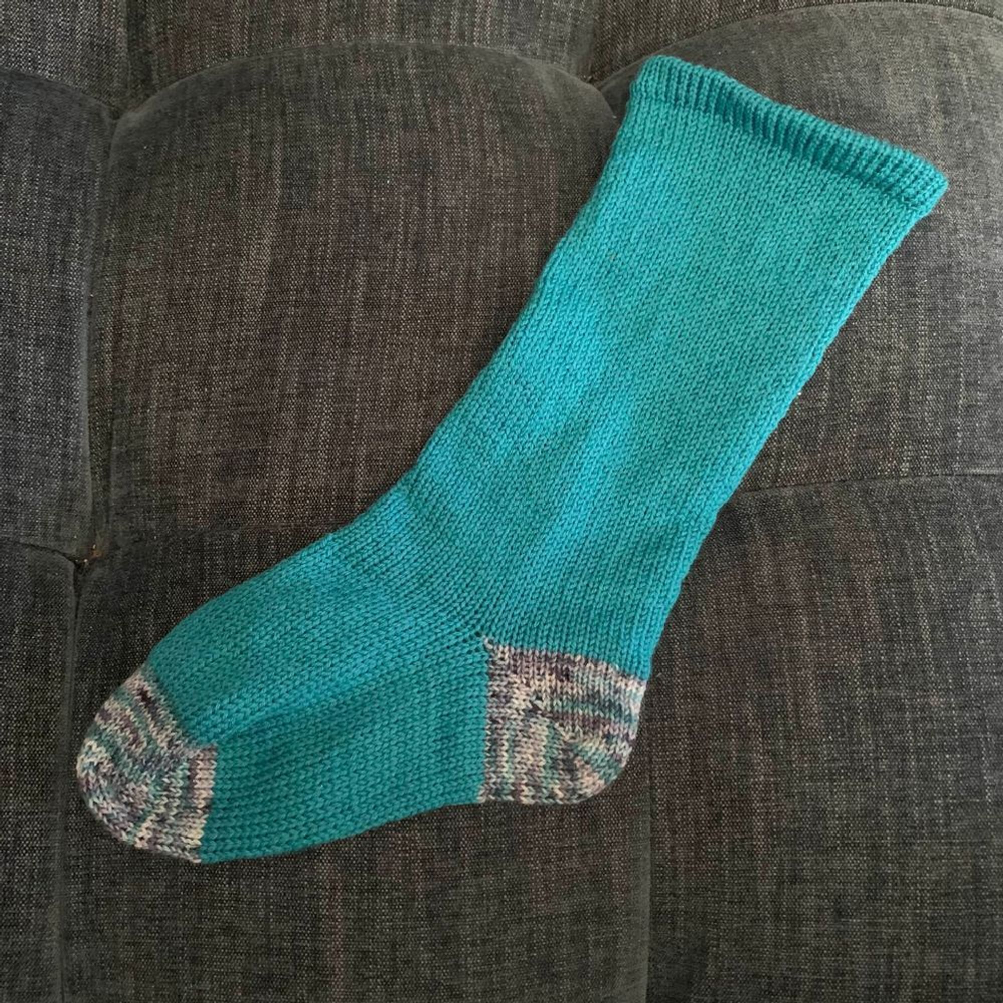 Photo of a knitted sock. The sock is primarily teal in color and has varigated toes and heel.