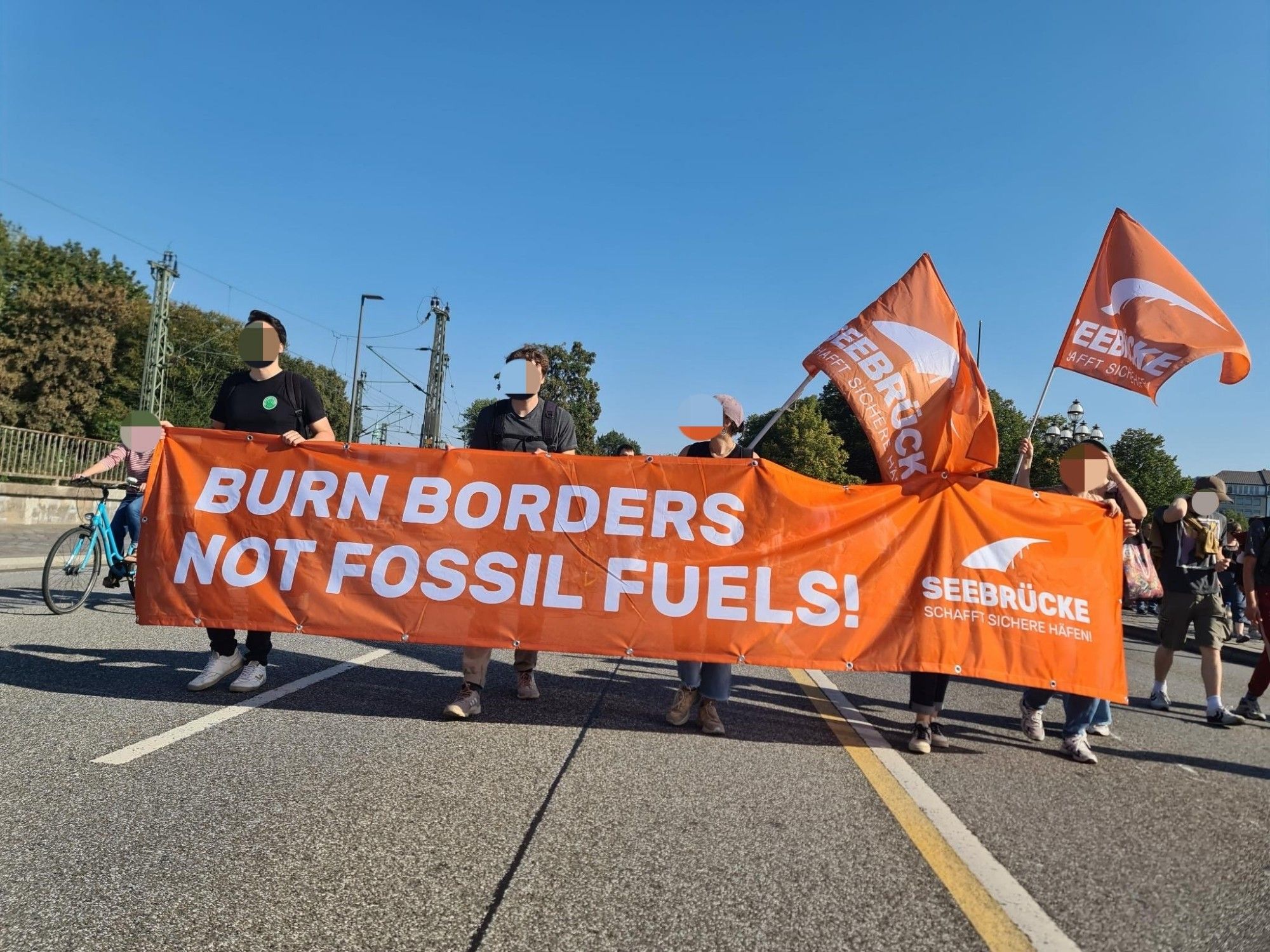 Foto von mehreren Menschen, die ein oranges Banner halten, auf dem "Burn borders Not Fossil fuels!" Steht. 2 Seebrücke-Fahnen werden dahinter getragen.