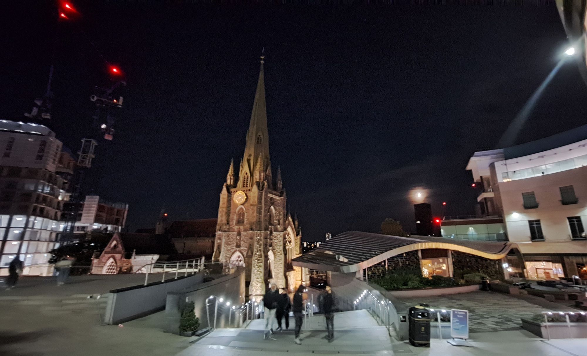 Night photo showing a jumble of architecture. From left to right: building under construction with cranes topped with red lights, English neogothic church mostly visible behind modern staircase with lit railings, a swoopy curved low roof (very nice Thai restaurant), distant ominous black tower with the moon above (very goth), and modern shopping centre on the far right