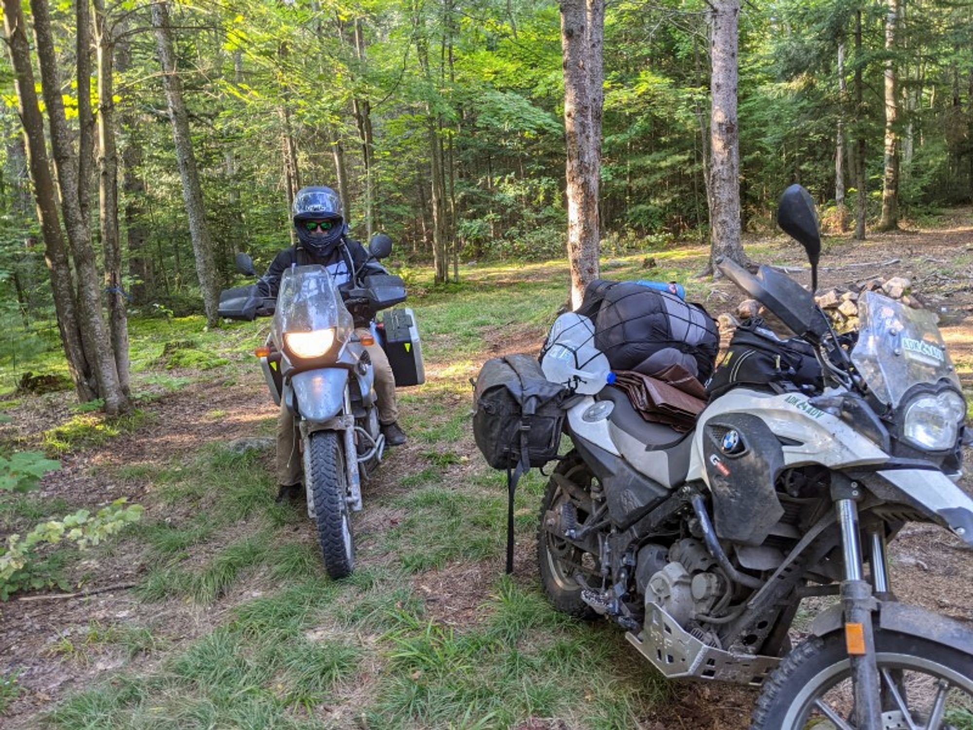 Two Adventure Motorcycles heavily loaded with supplies. One is riderless and the other has a helmeted rider. Sleeping bags, water, tarp are visible strapped on to overstuffed saddlebags.