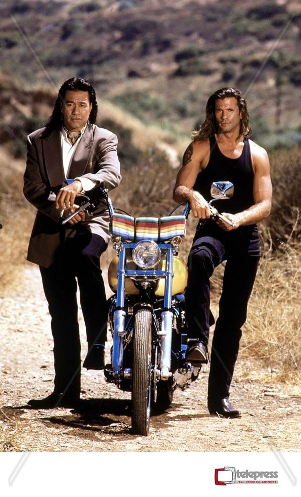 Promotional image for TV Show Renegade. Two characters, one standing on each side of a motorcycle. The background is Southwest dessert.