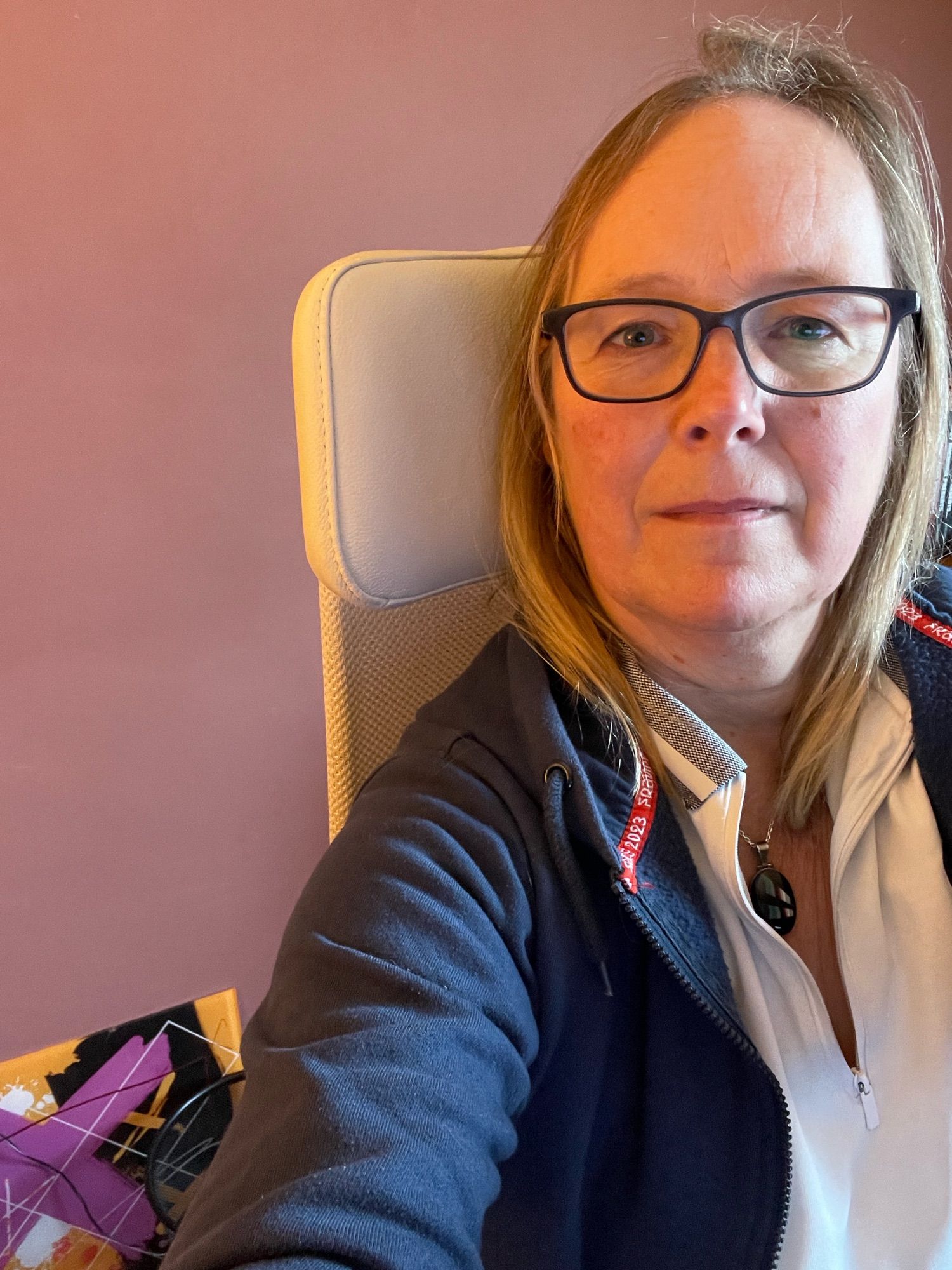 A selfie of a bespectacled blonde woman with the utter ergonomic beauty of a headrest visible behind her. Glimpse of an abstract artwork in the corner of the picture.