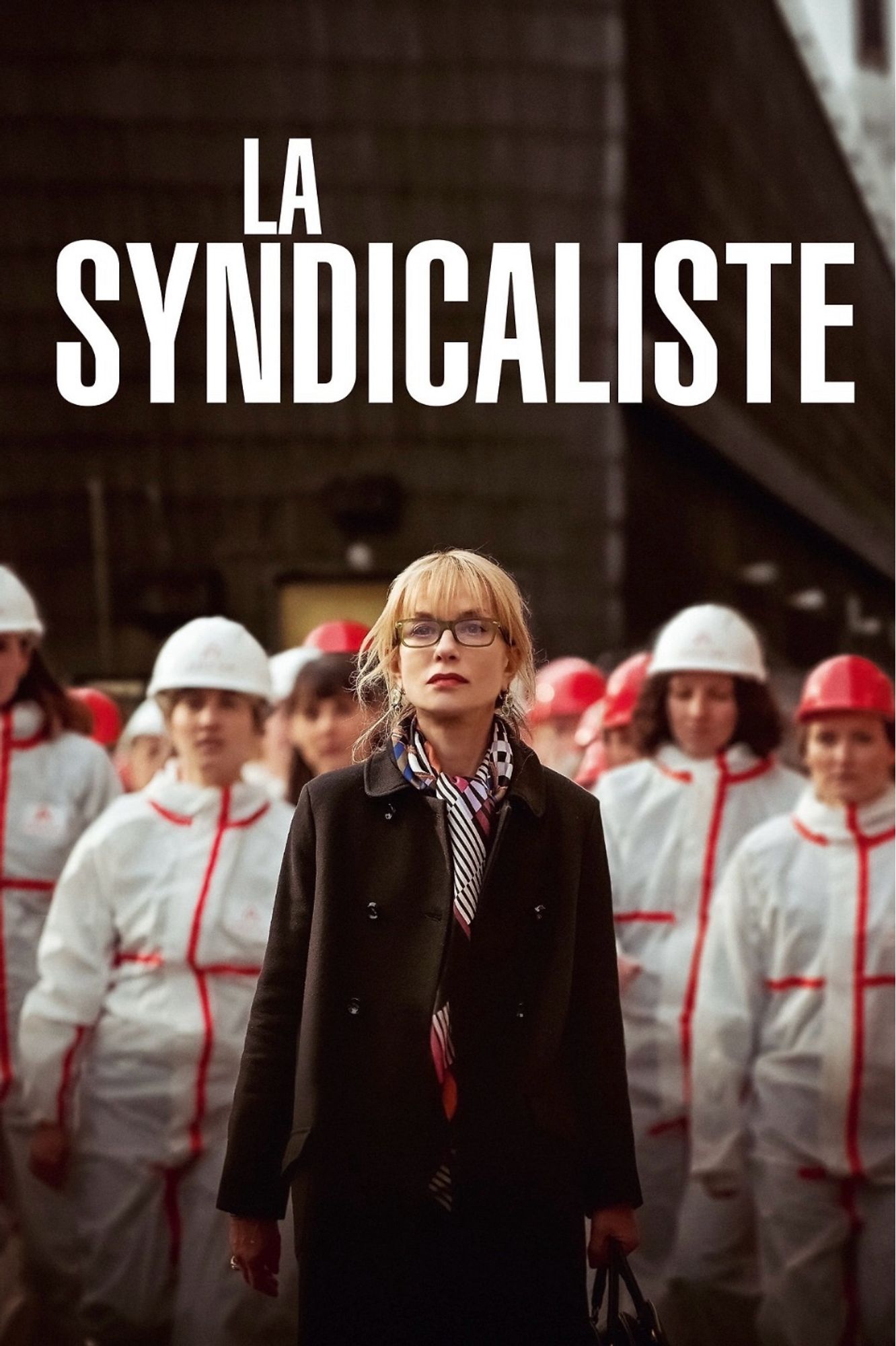 Poster for La Syndicaliste. The poster shows Isabelle Huppert in a dark jacket and glasses, witha  group of woman in white work overalls and hard hats standing behind her in front of a factory.