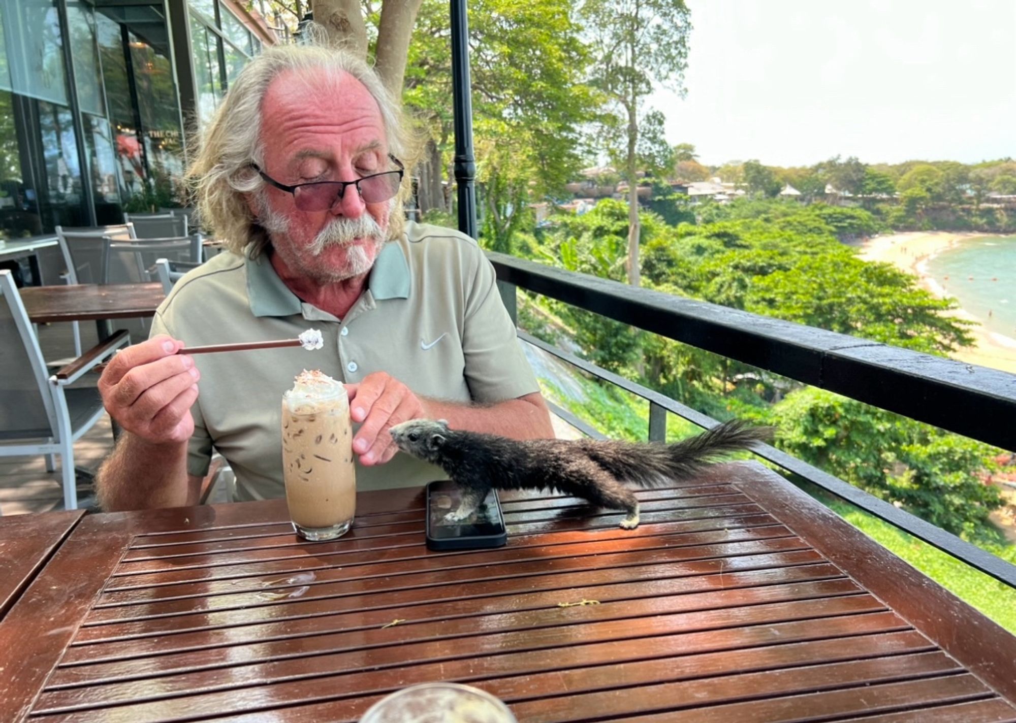 My dad, a white man with long hair, drinking a once coffee. His mobile phone is on the table, and there is a brown squirrel with a grey face standing on his phone, sniffing at the coffee