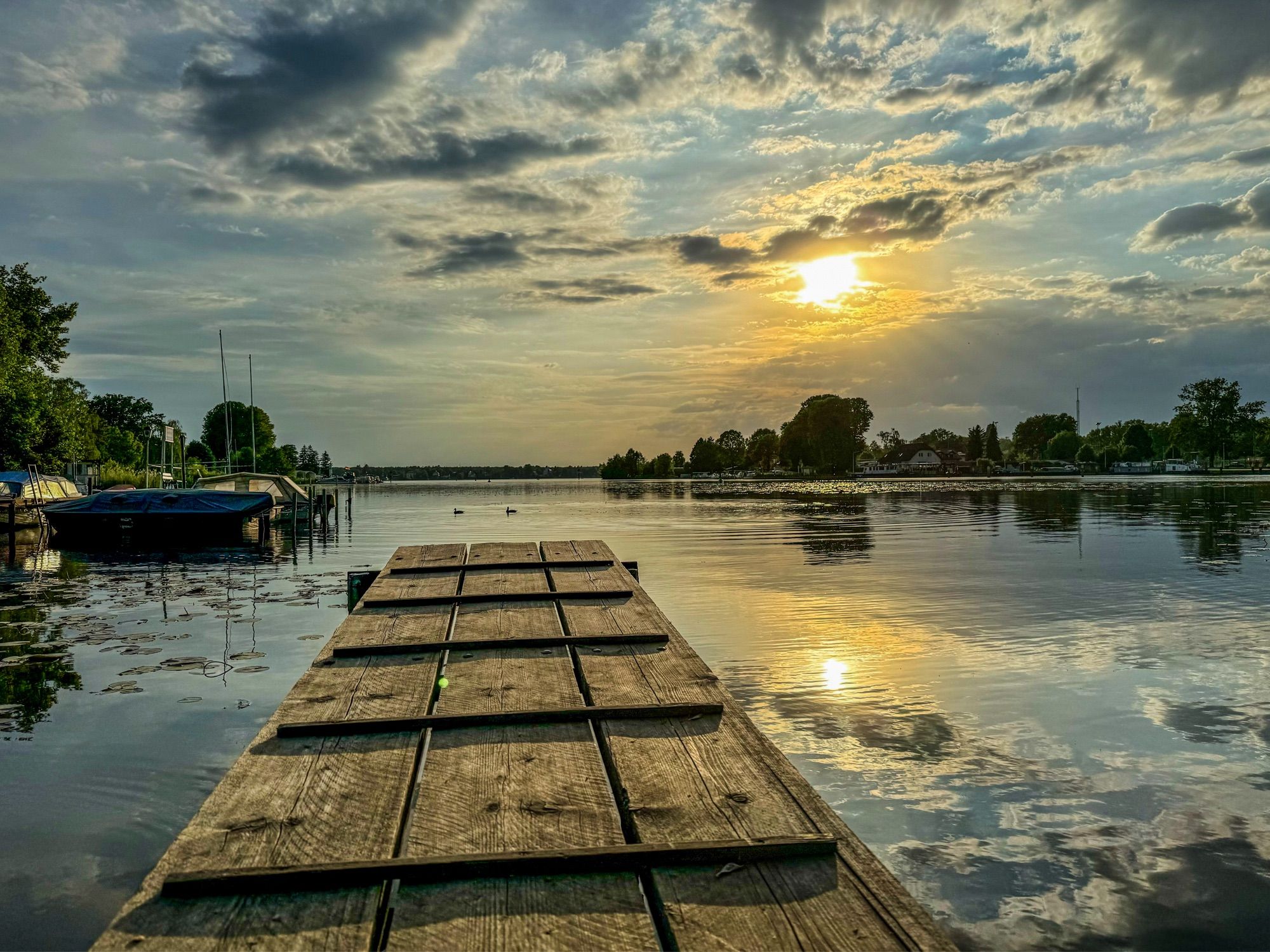 See mit Steg, Sonne tendiert Richtung Untergehen und spiegelt sich gemeinsam mit Wolken im Wasser.