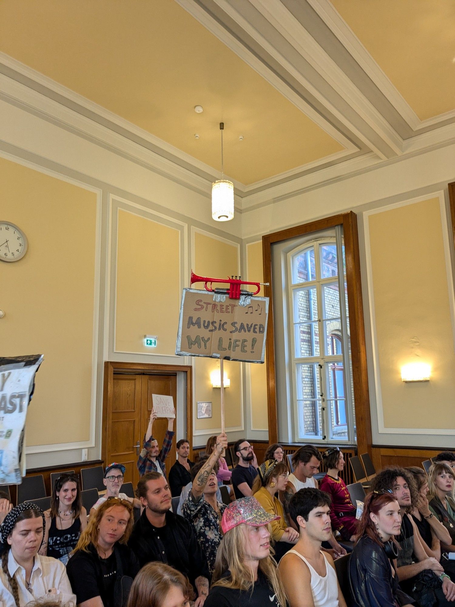 Besucher*innen im Saal der Bezirksverordnetenversammlung Pankow. Ein Transparent "Street music saved my life!" wird hochgehalten.