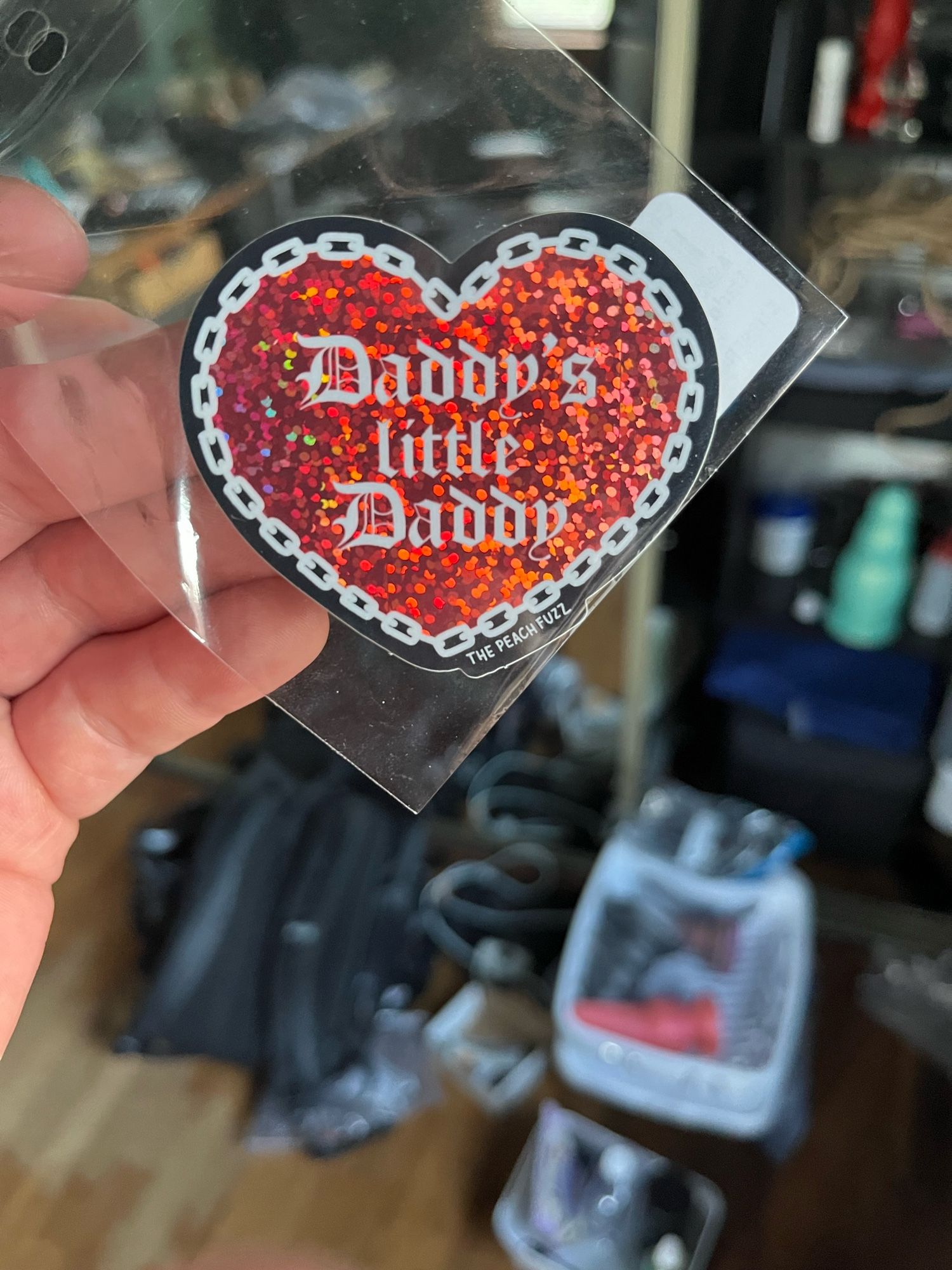 Red, holographic heart with a chain border. It reads “Daddy’s little Daddy” in old english lettering from top to bottom.