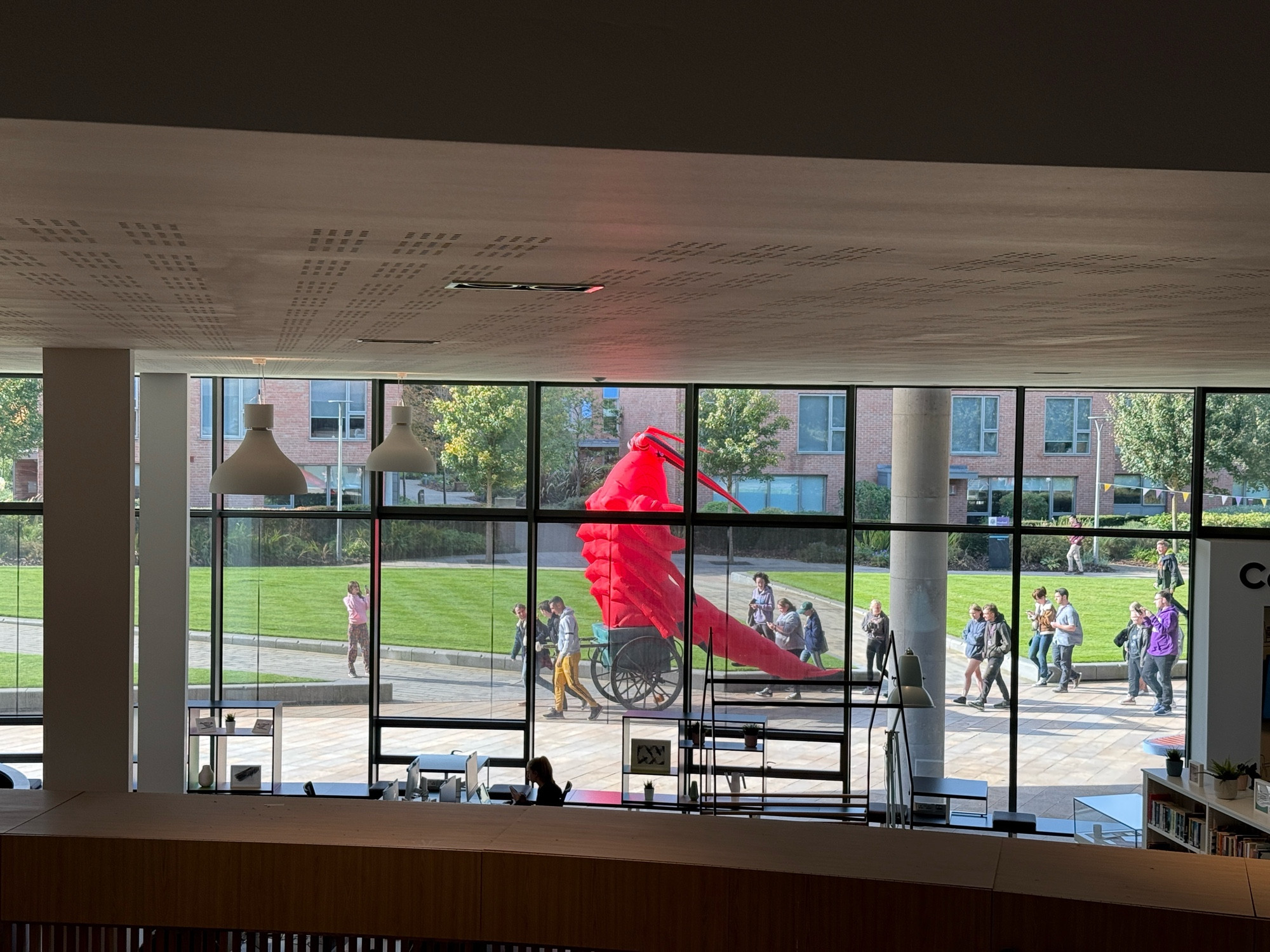 A photograph through a window of a giant inflatable lobster on a trolly being followed by a group of students.