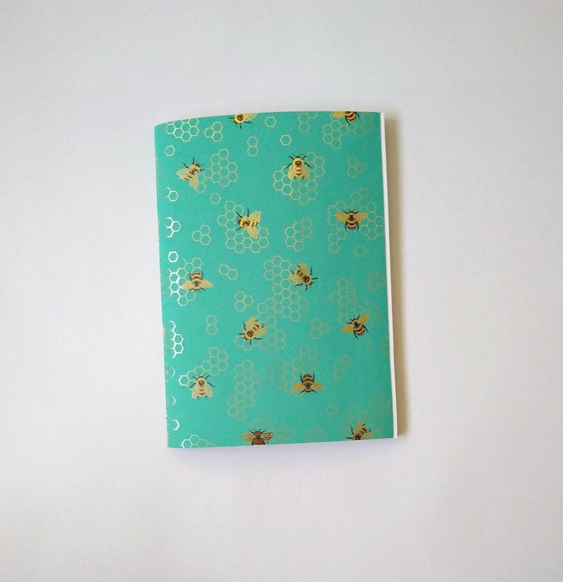 A photograph of a green A5 sketchbook sketchbook against a white background. The book is patterned with golden bees and honeycomb pattern.