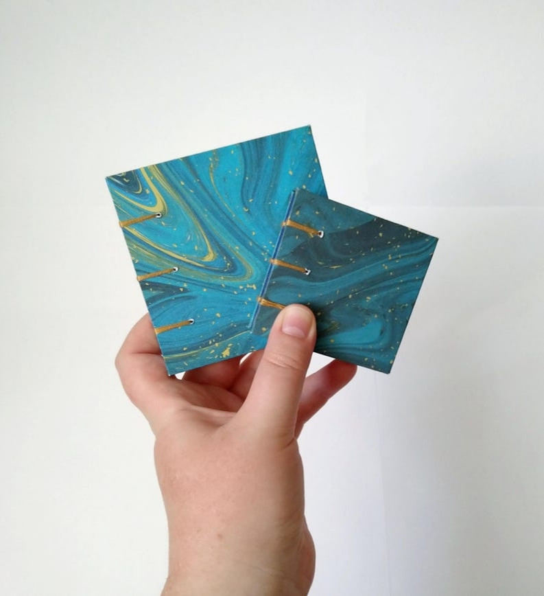 A photograph of a hand holding two small blue and gold sketchbooks, both smaller than the hand.