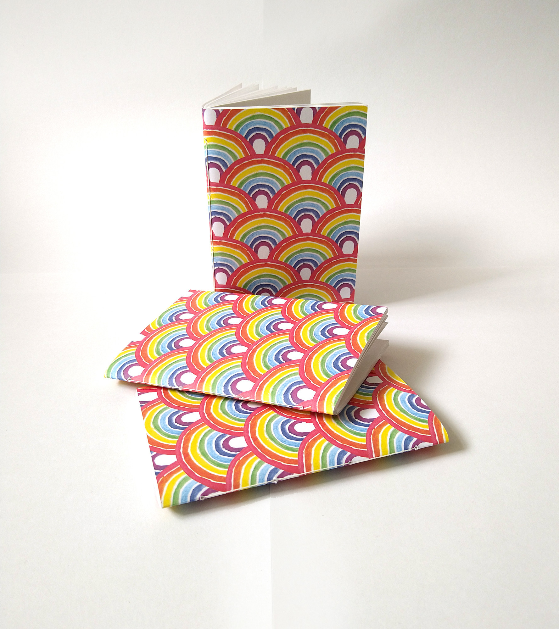 a photograph of three A6 paperback notebooks against a white background. Two are lying down and one is standing up on its end. They are all patterned all over with a repeating watercolour rainbow arch pattern.