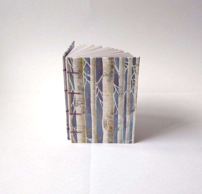 A photograph of a blue sketchbook standing up against a white background. The sketchbook is patterned with illustrated silver birch trees against a blue and gold background.