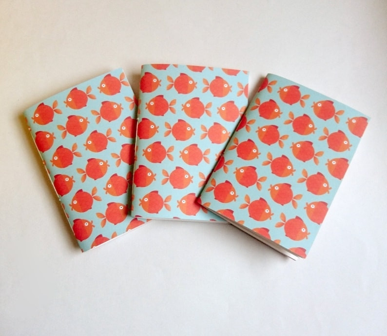 A photograph of three paperback A6 sketchbooks on a white background. The sketchbooks are pale blue and patterned with stylised orange goldfish.