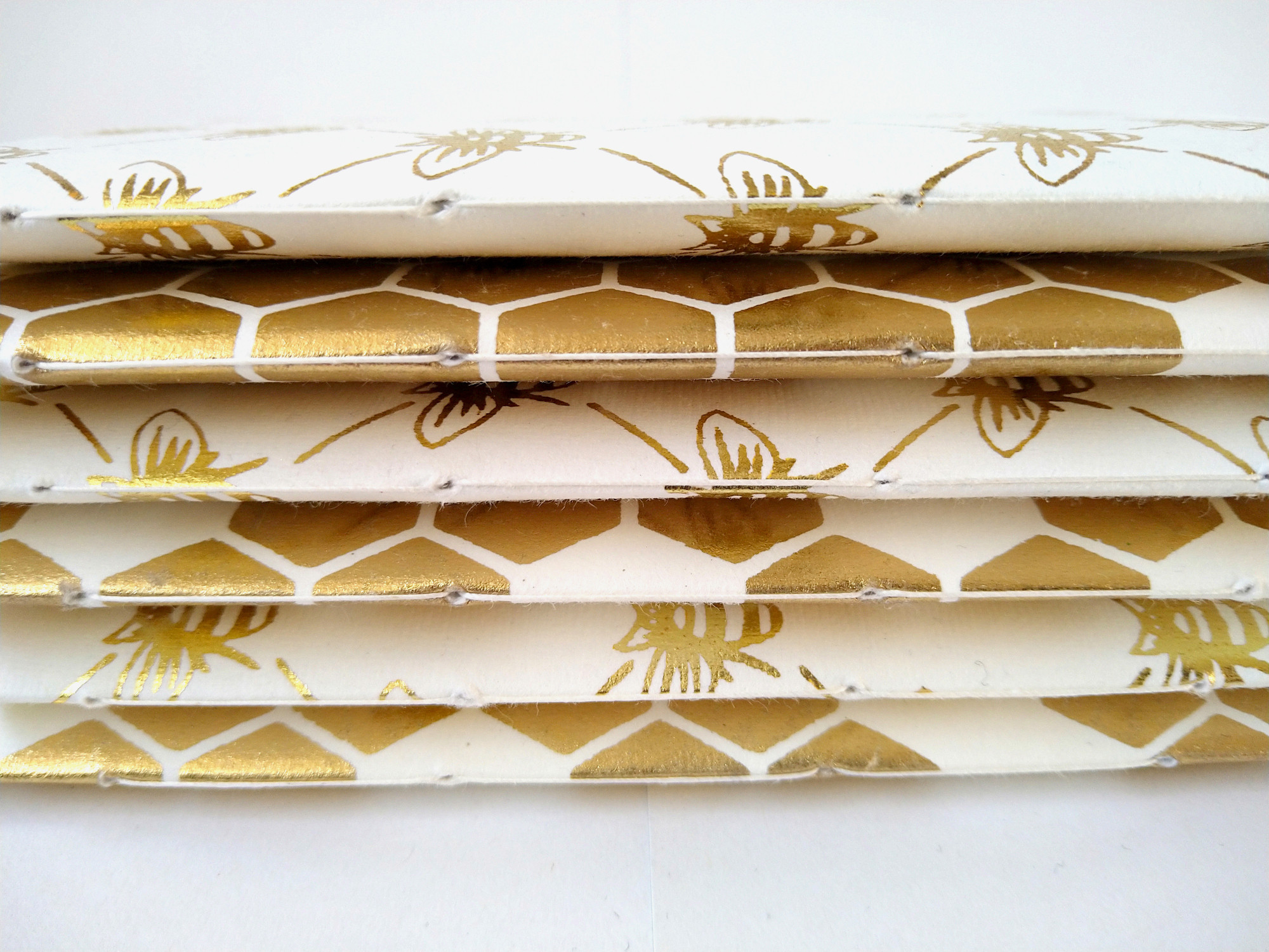 A photograph of the spines of several paperback A6 notebooks in a stack, on a white background. The notebooks are alternately patterned with gold bees, or gold honeycomb.