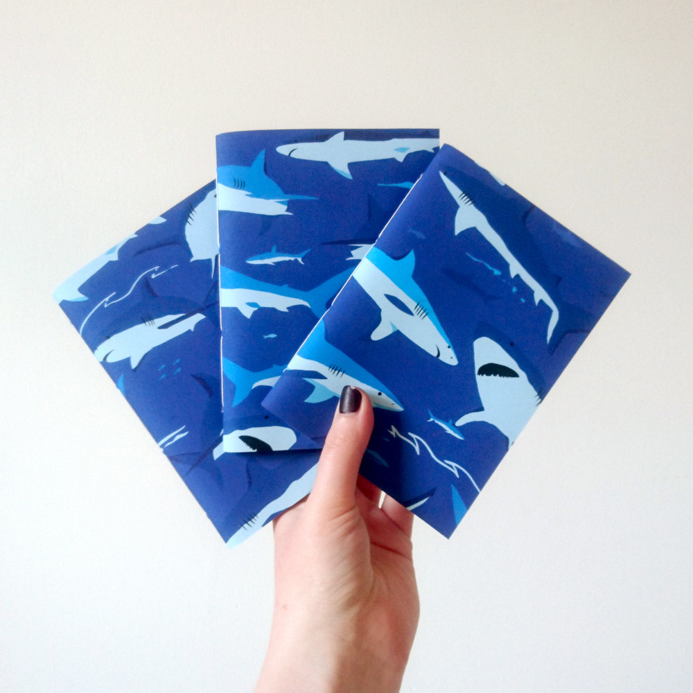 A photograph of three A6 paperback notebooks held up in a fan by a white hand against a white background. The notebooks are dark blue and patterned with illustrated sharks in various shades of blue.