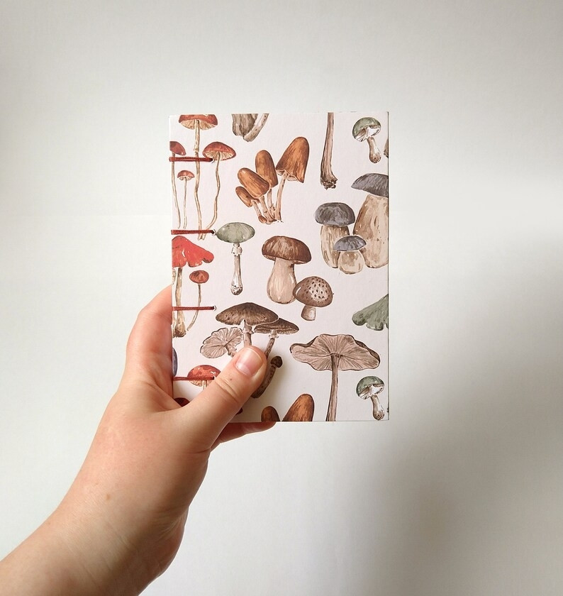 A photograph of a hand holding an A6 sketchbook against a white background. The sketchbook is white and patterned with illustrated mushrooms.