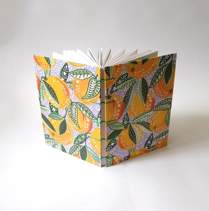 A photograph of a yellow, green and purple sketchbook standing up open, against a white background. It is patterned with oranges and has a Coptic bound spine.