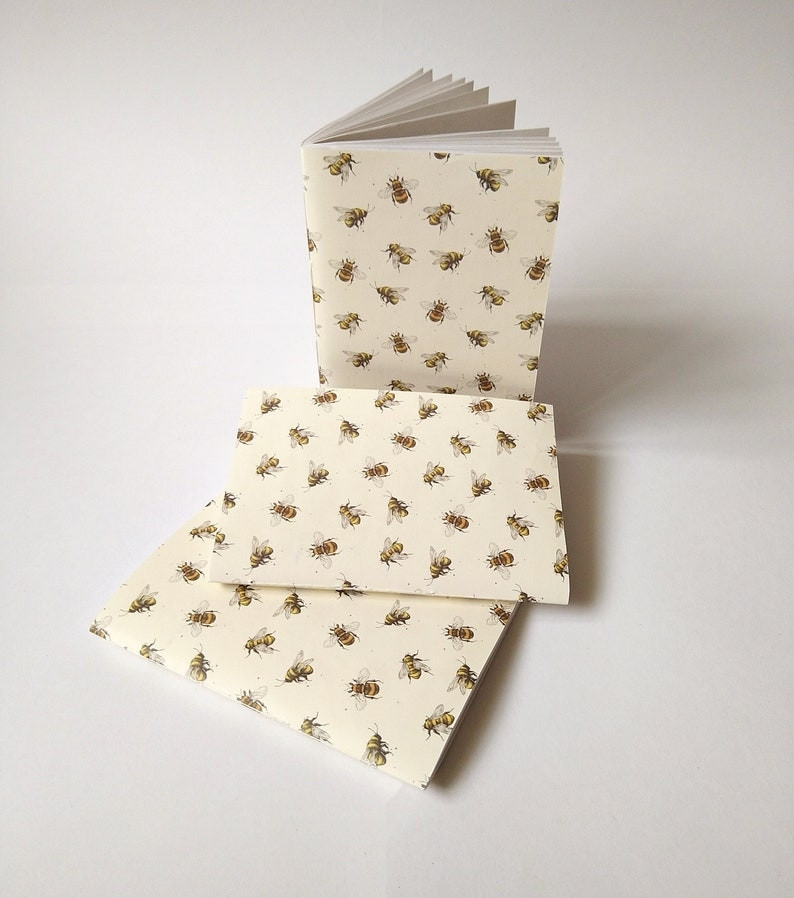 A photograph of three A6 sketchbooks against a white background. The sketchbooks are cream coloured and patterned with small illustrated bees.